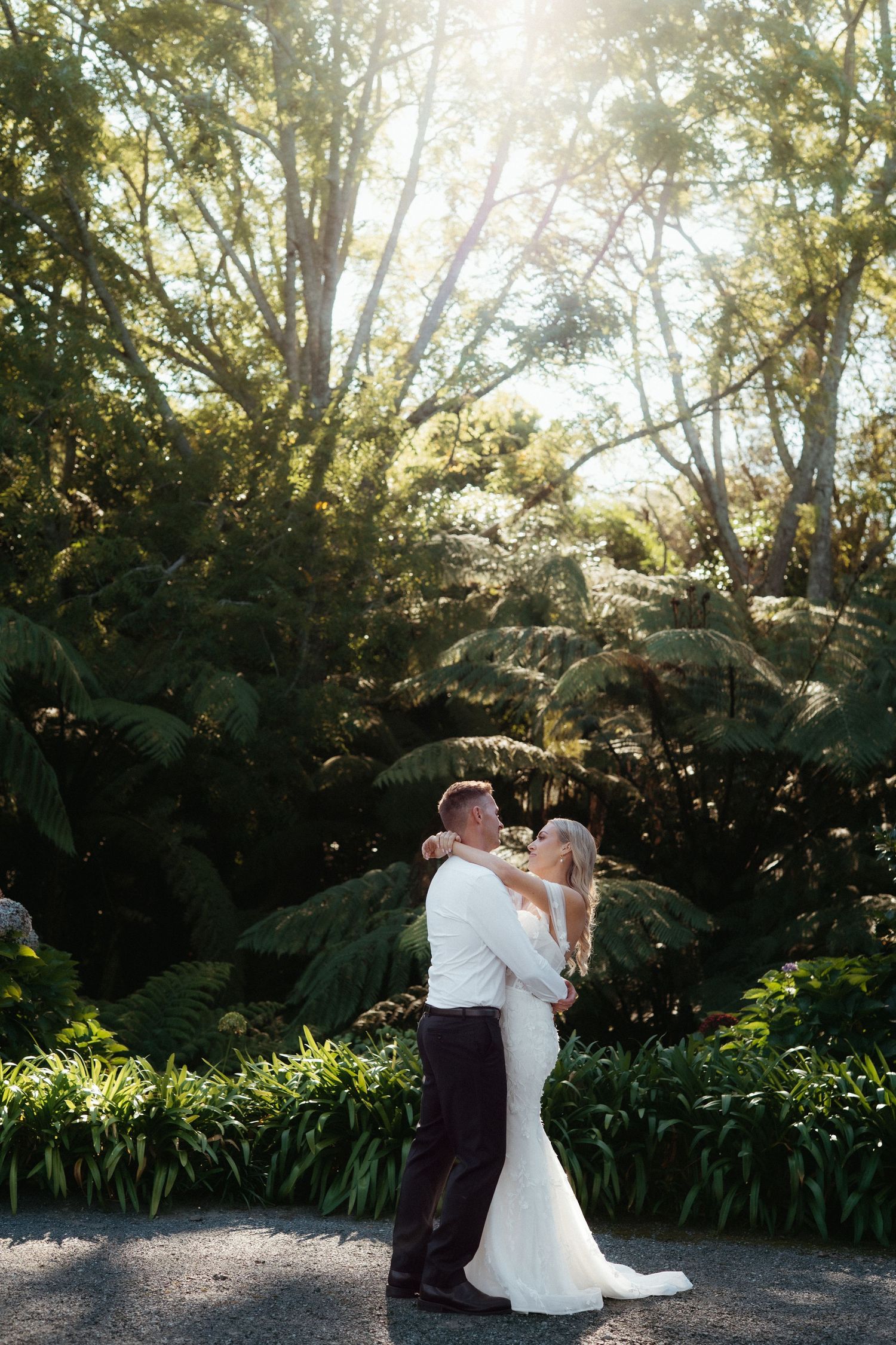 Bride and groom embracing in golden sunlight surrounded by lush tropical ferns and trees.