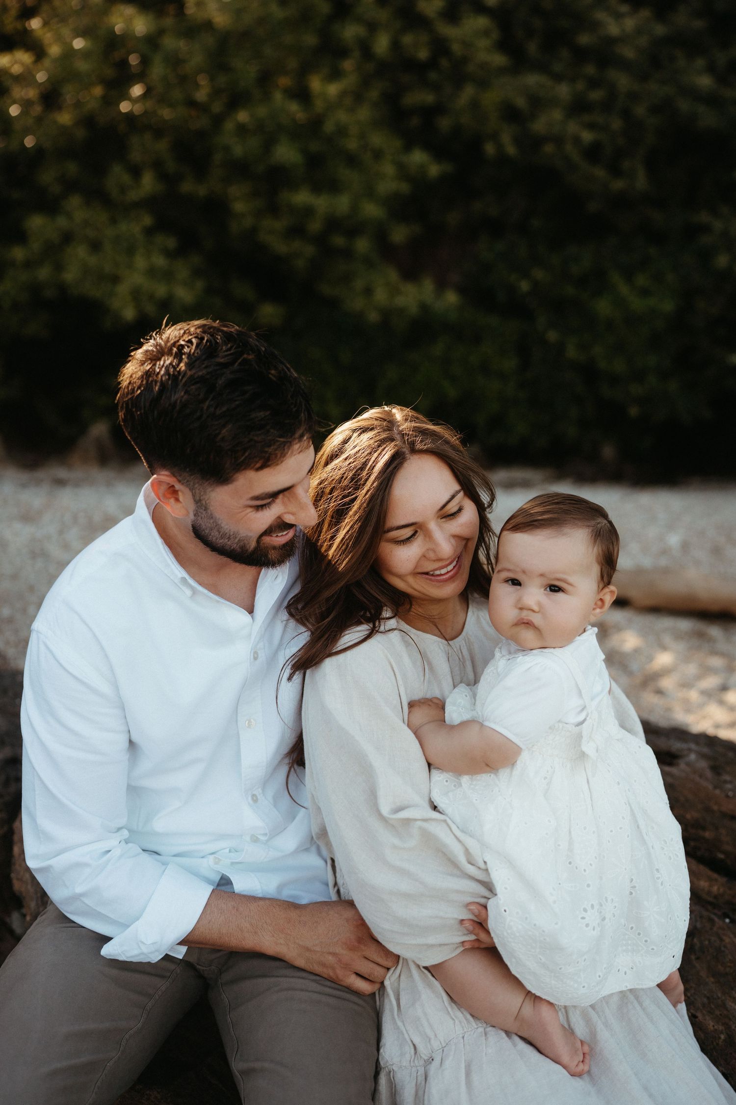 Happy couple sitting outdoors holding their baby girl dressed in white in warm golden light.