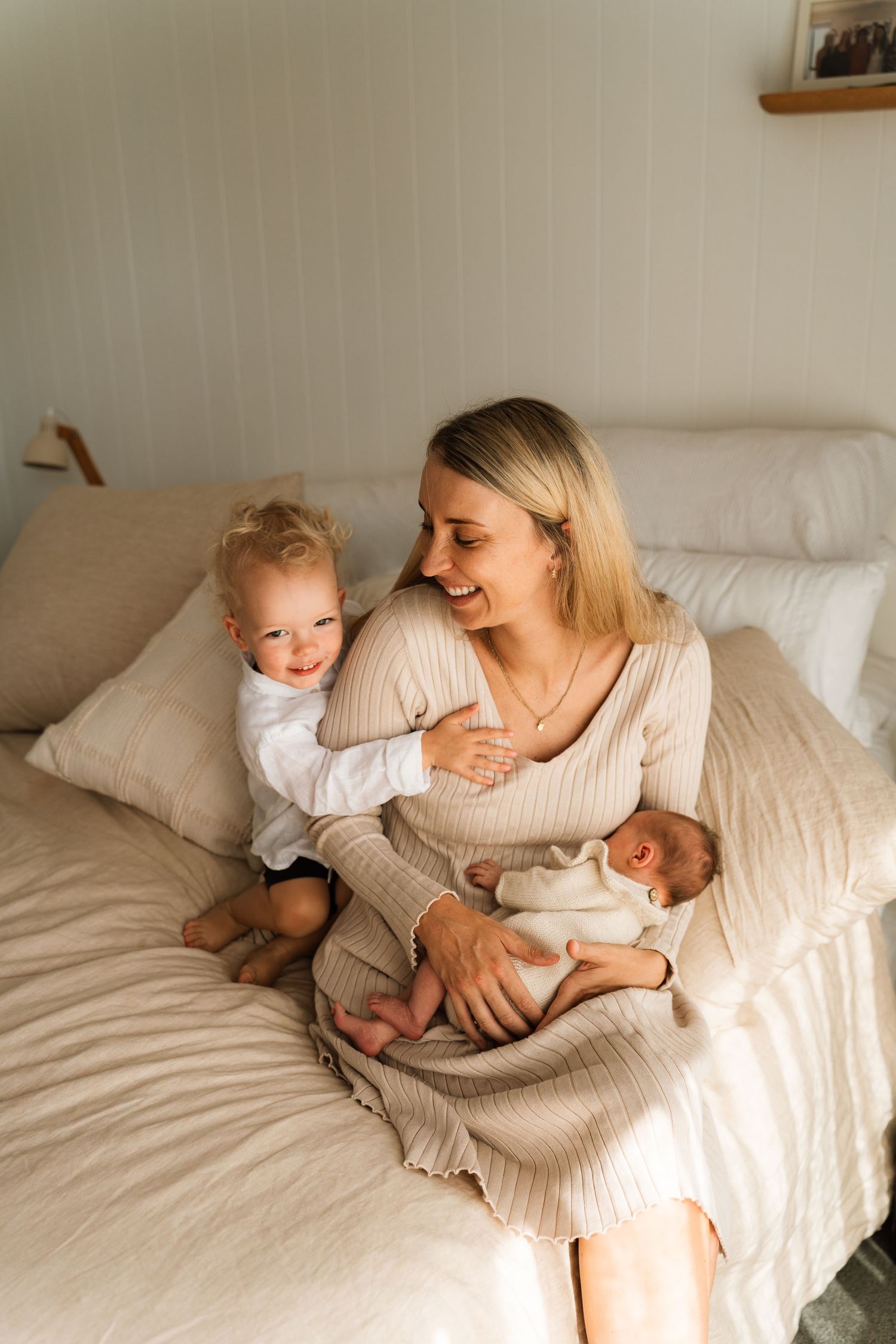 A smiling mother sits on a bed holding a newborn while her toddler hugs her in a cozy bedroom.