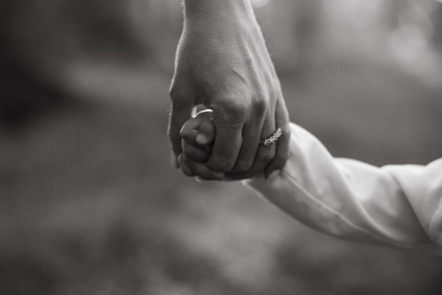 Black and white close-up of parents holding a small childs hand with wedding rings visible.