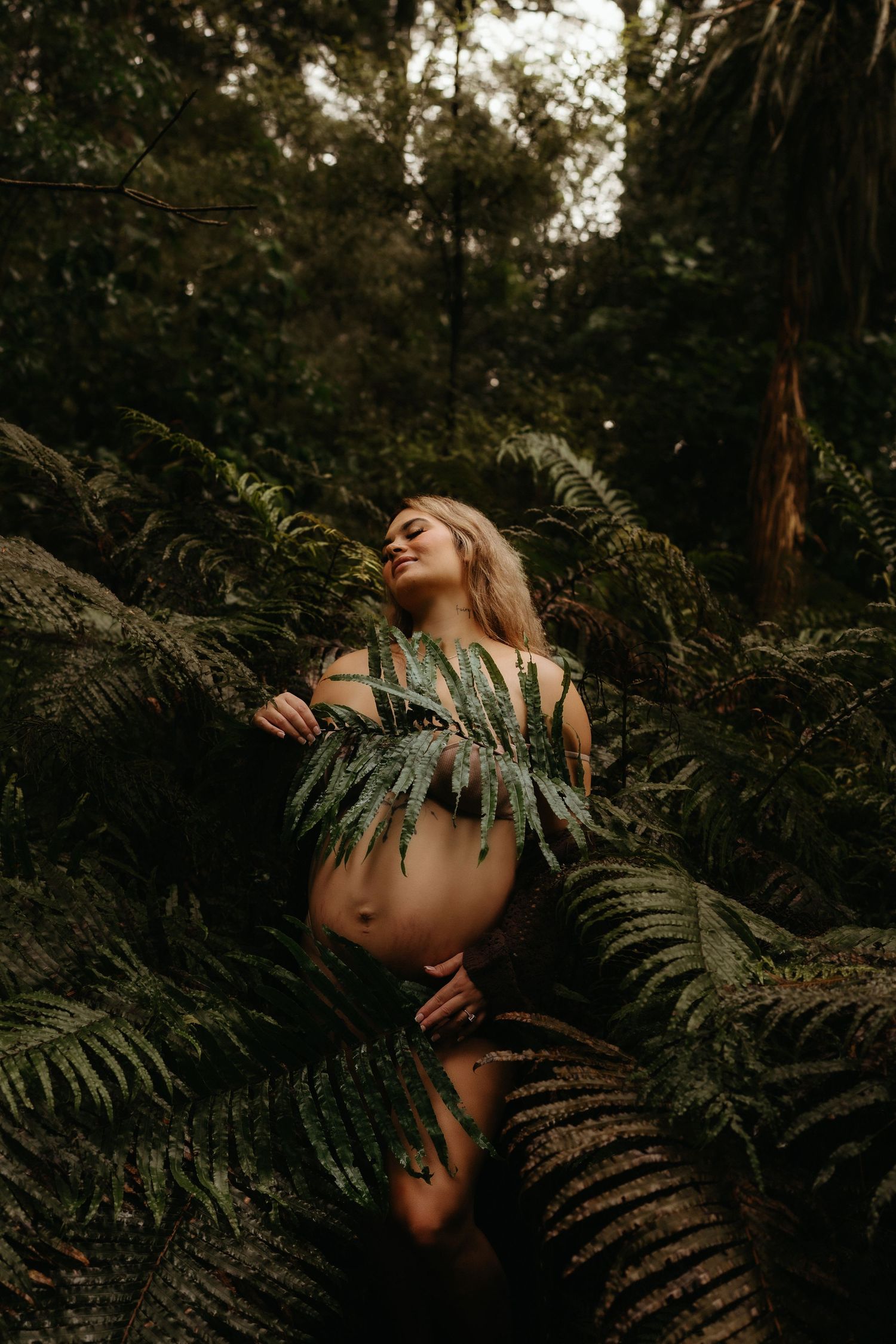 Pregnant woman posing nude among lush green ferns in a dark forest maternity shoot.