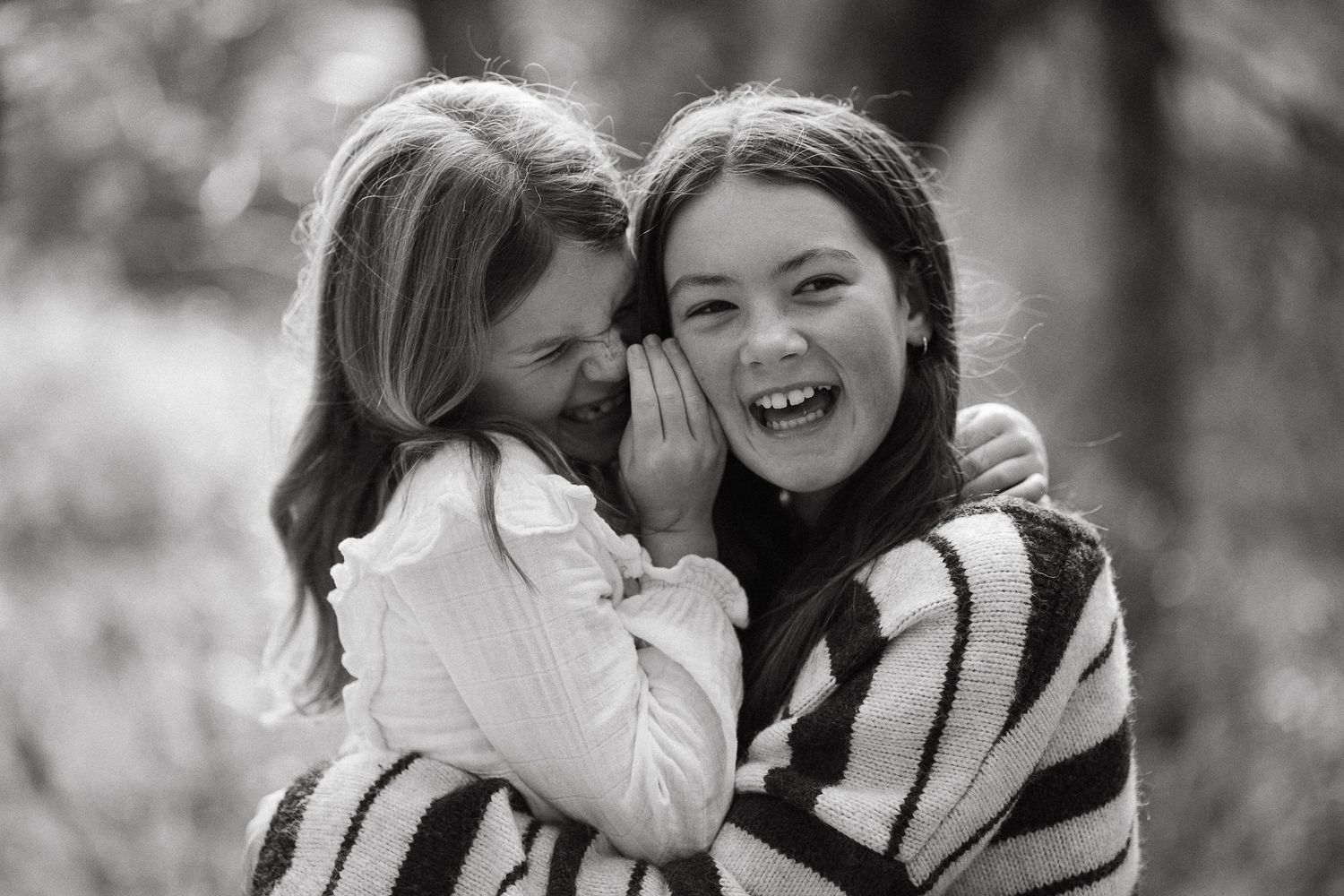 Two girls laughing and sharing a secret outdoors in a black and white photo.