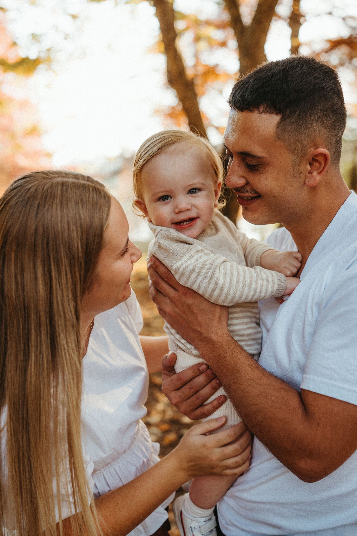 Smiling baby held by parents in a warm autumn outdoor setting.