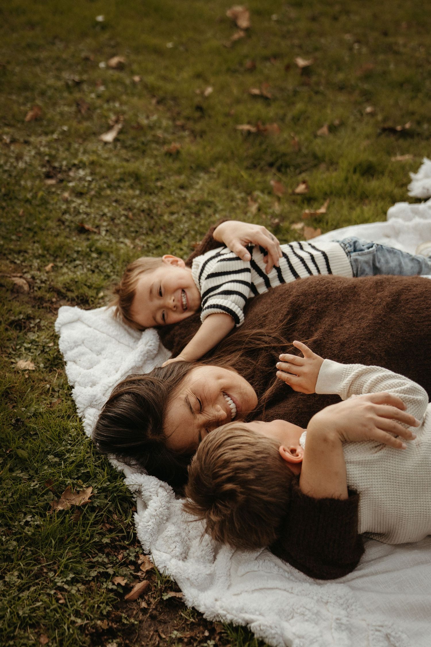 A mother and two young children lying on a white blanket on green grass, smiling and cuddling together outdoors.