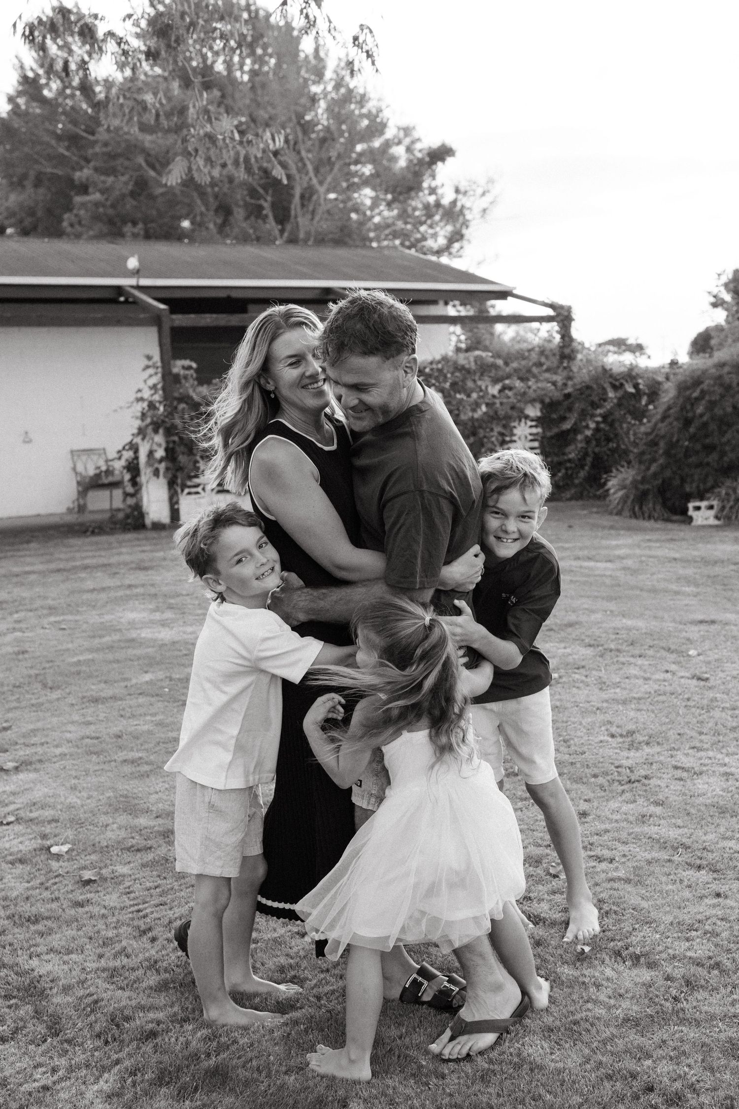 Black and white photo of a happy family with three children hugging their parents in a backyard.