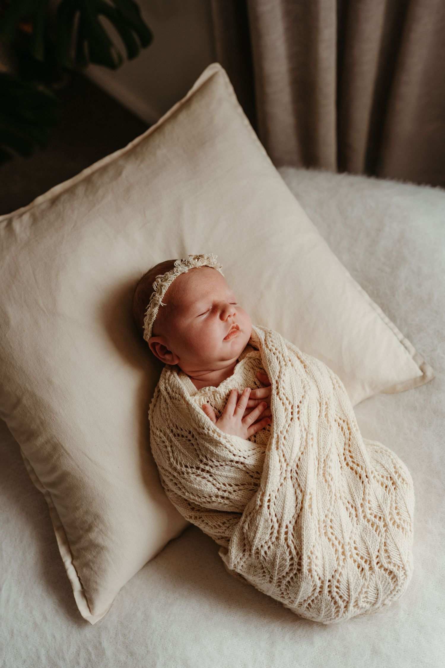 Newborn baby swaddled in cream knit wrap with lace headband, sleeping peacefully on beige pillow.