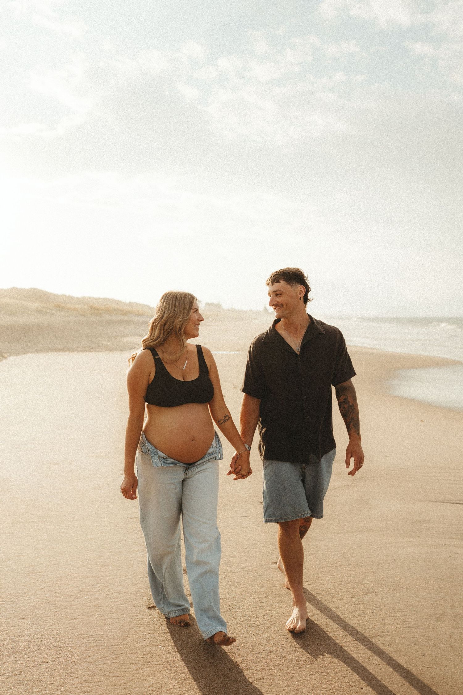 Pregnant couple holding hands and walking along a sunny beach during maternity photoshoot.
