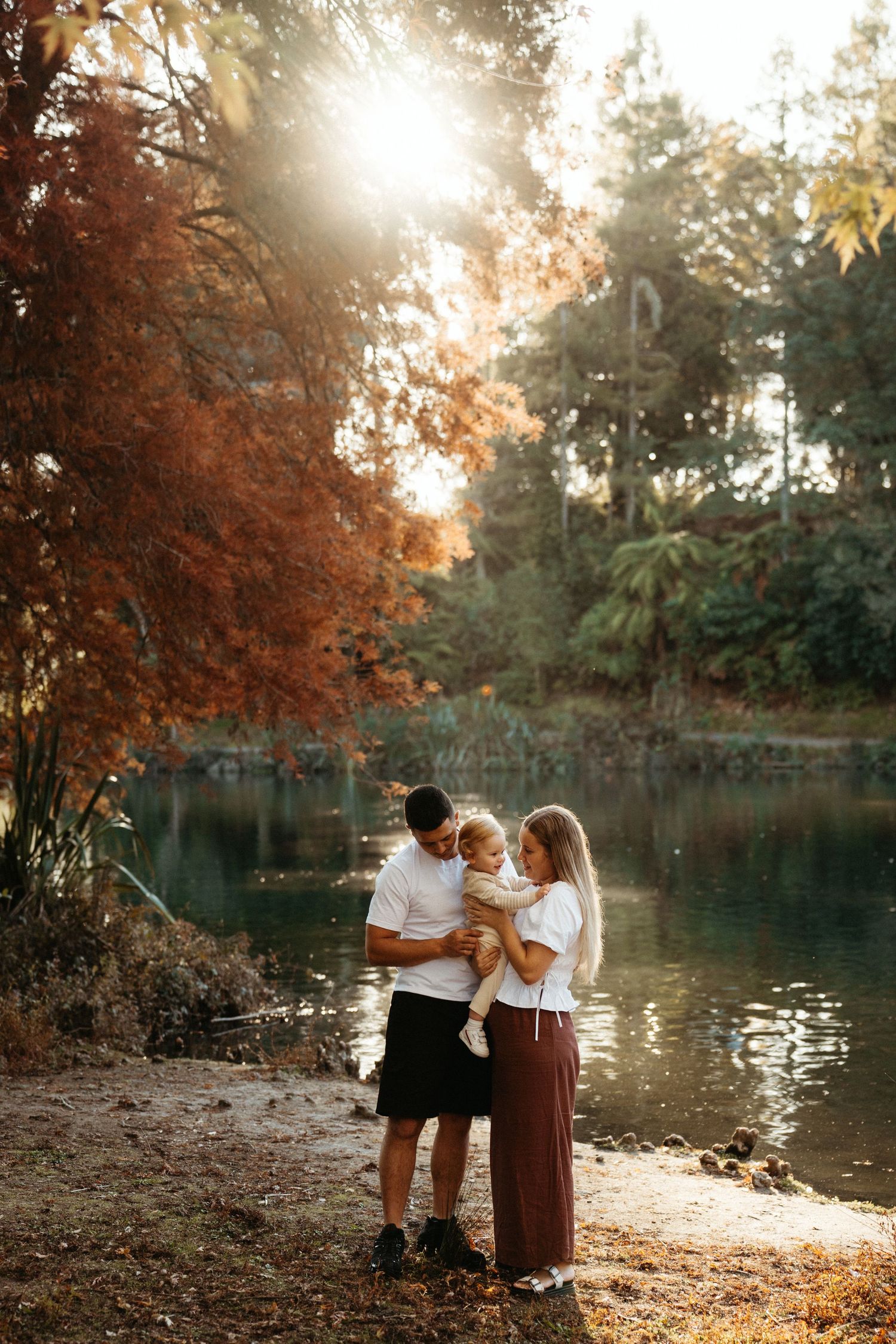 Young family with baby standing by a tranquil lake surrounded by colorful autumn trees.