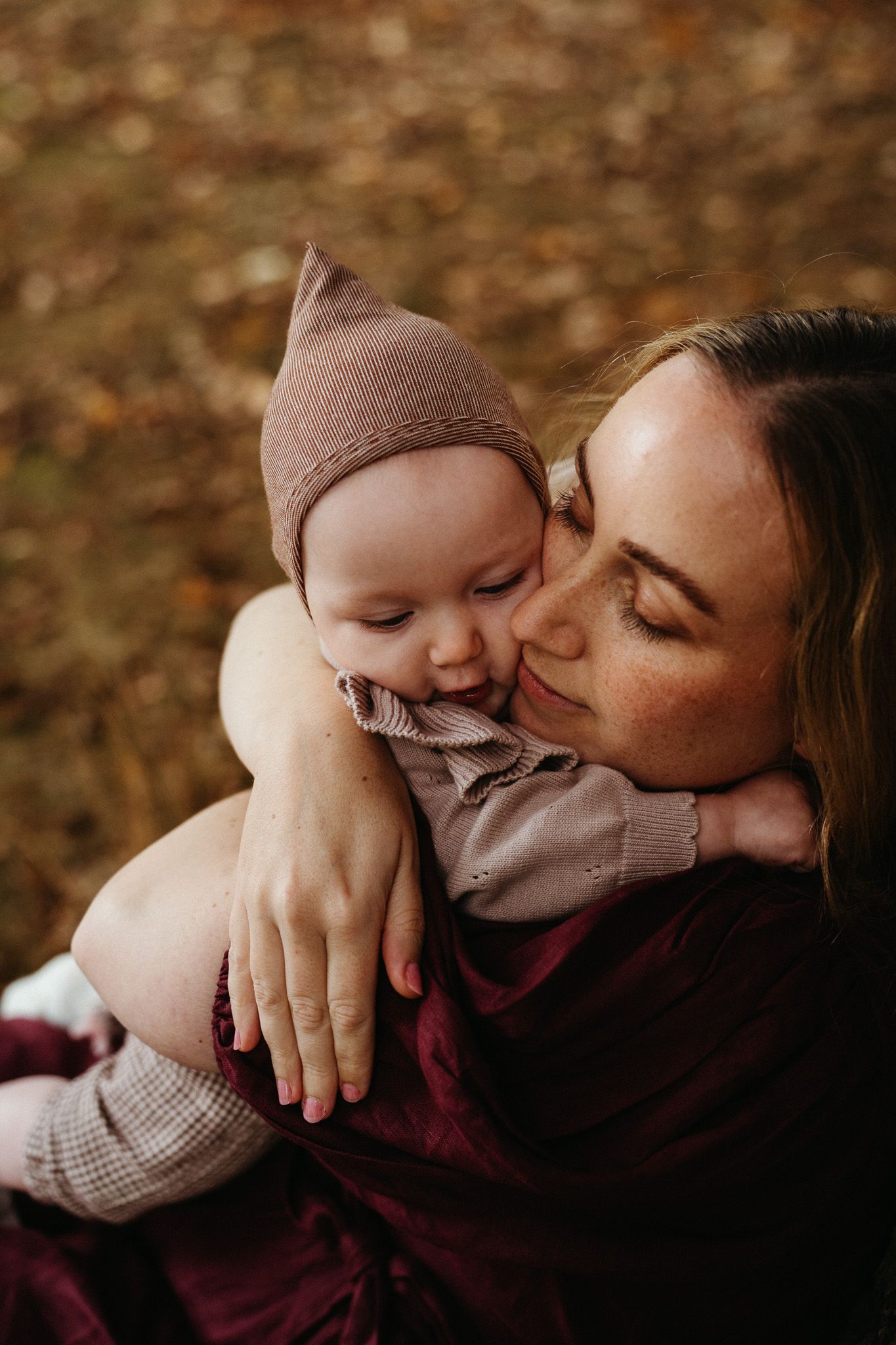 Mother with freckles tenderly kissing her baby wearing a pink bonnet in autumn leaves.