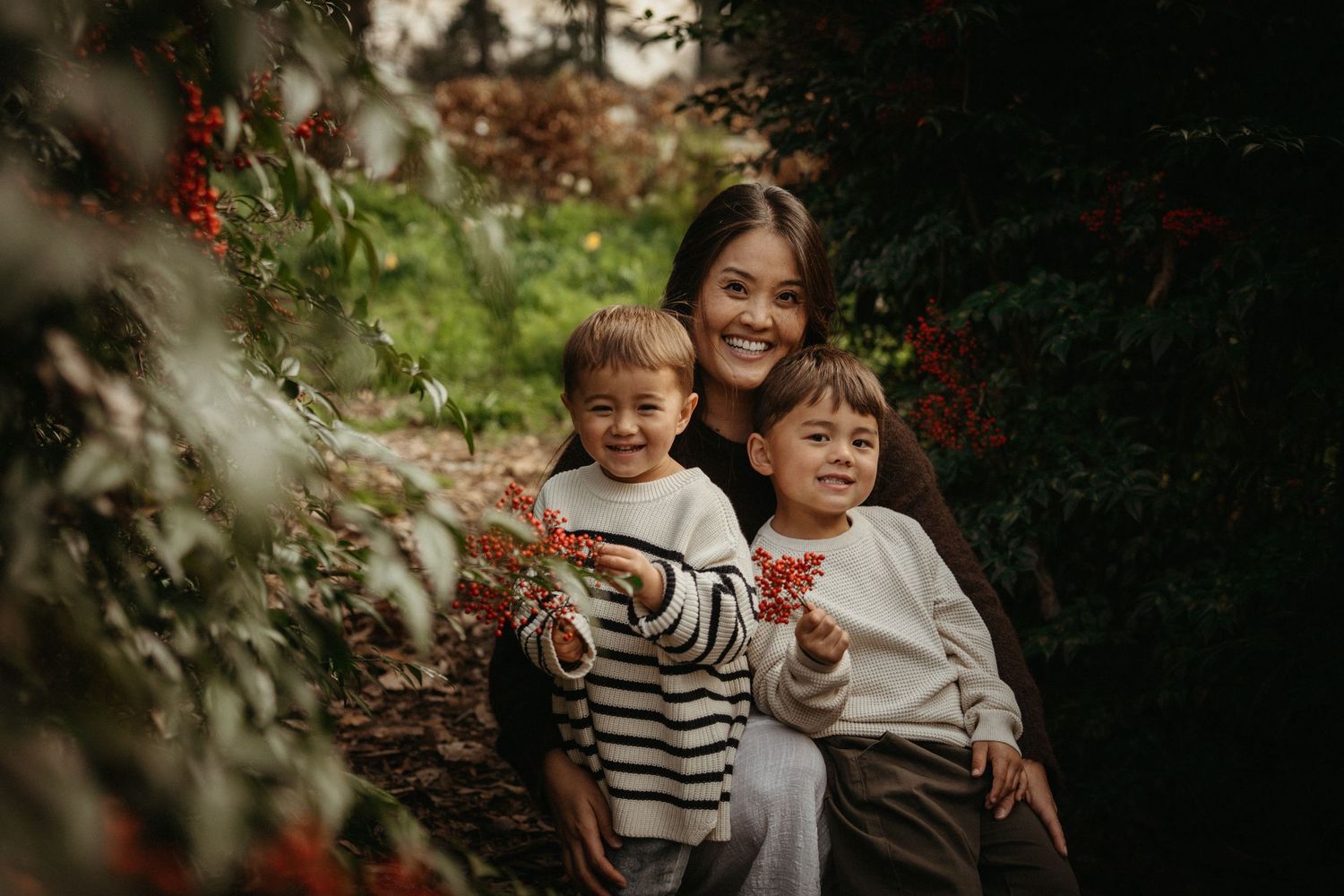 A smiling woman poses with two young children among lush green and red berry bushes in a garden setting.