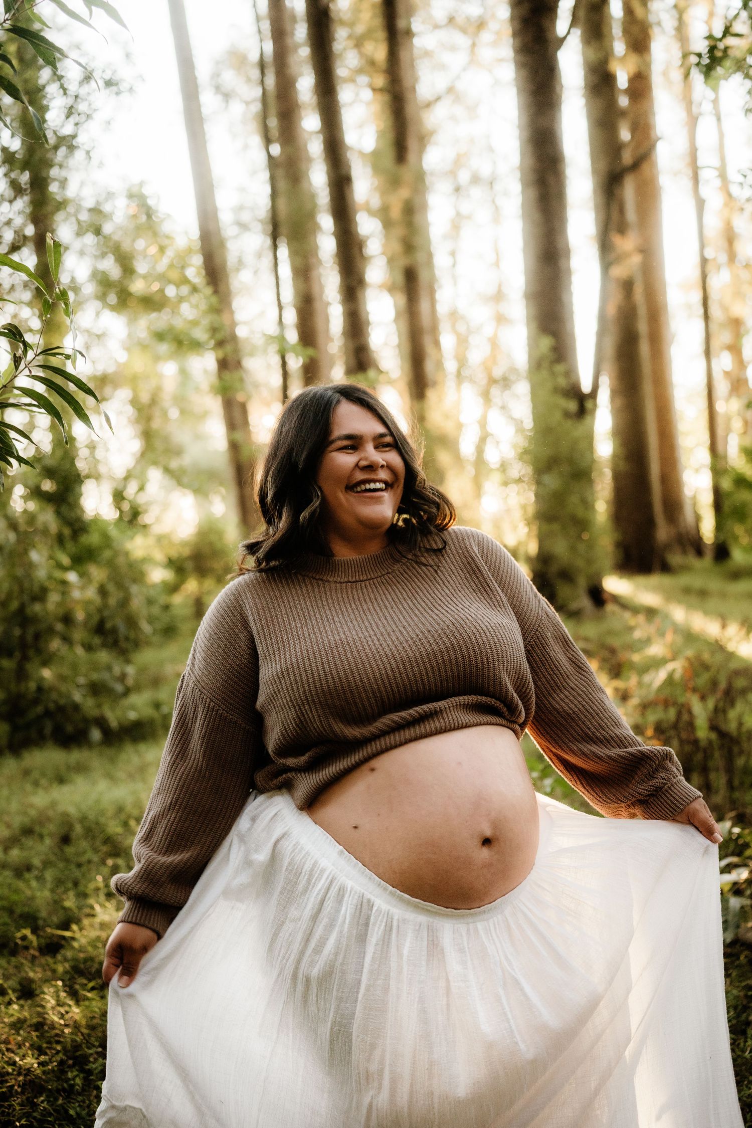 Smiling pregnant woman in brown sweater and white skirt posing outdoors in a sunlit forest.