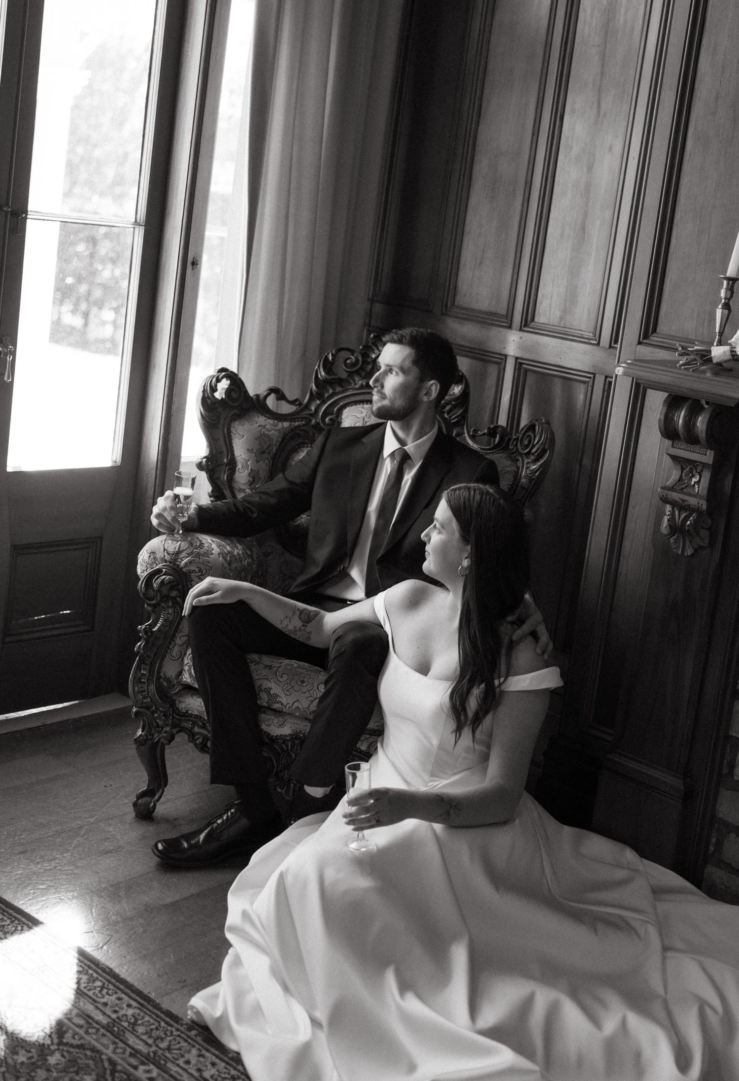 Bride and groom sit together in an ornate vintage chair gazing out a window in black and white.