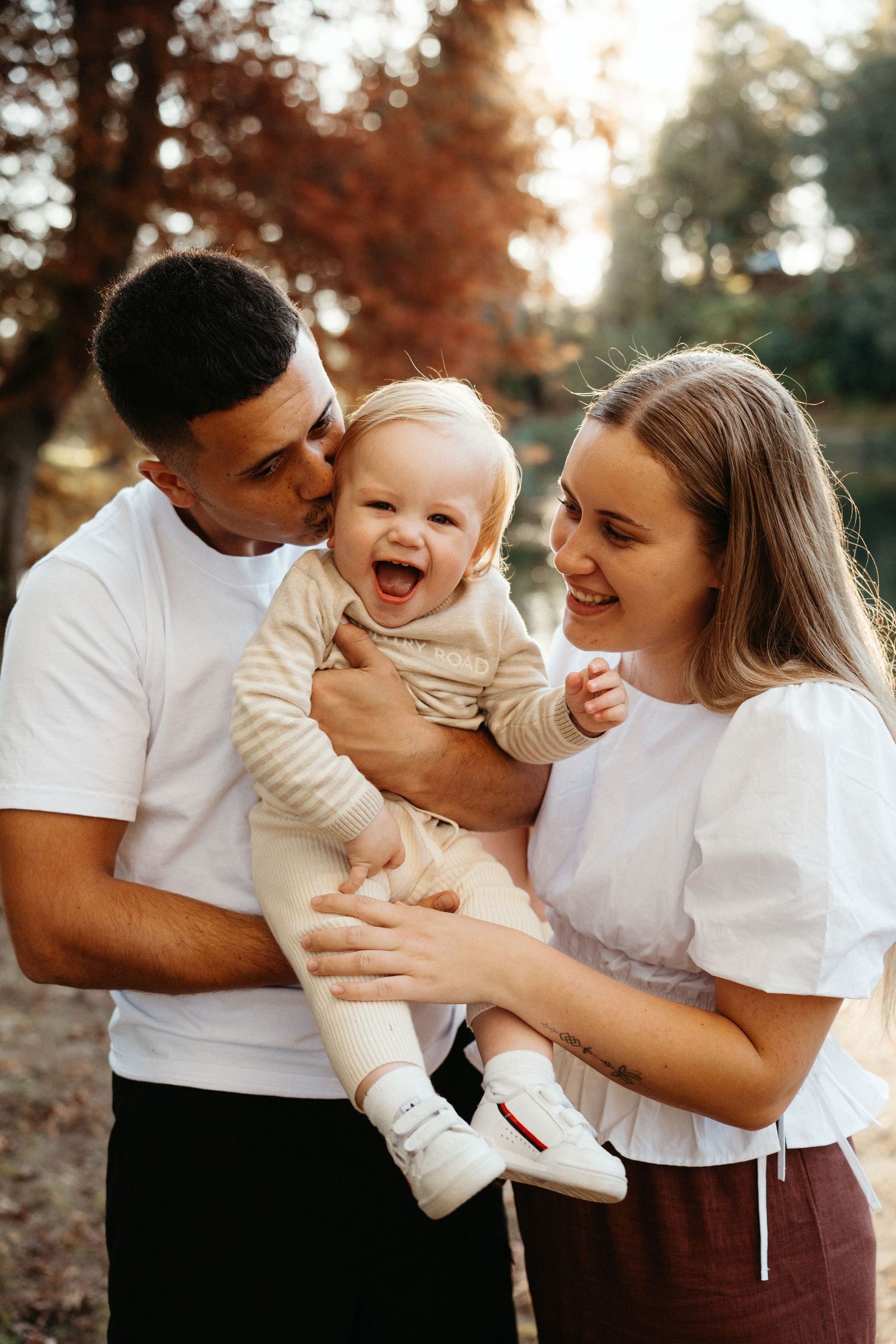Joyful baby laughing while being held by parents in a sunlit autumn park.
