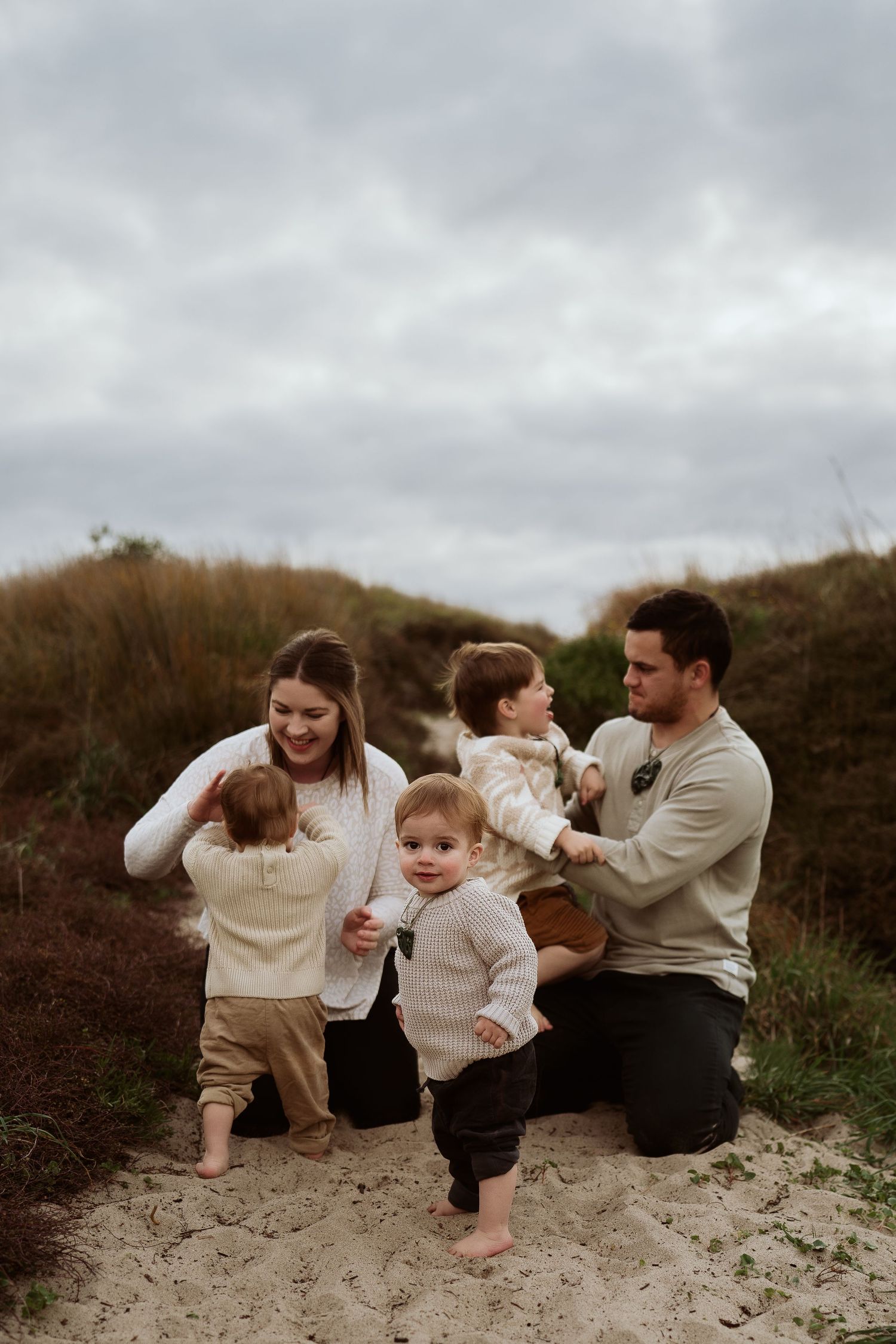 Young family with three toddlers playing together on sandy beach dunes under cloudy sky.