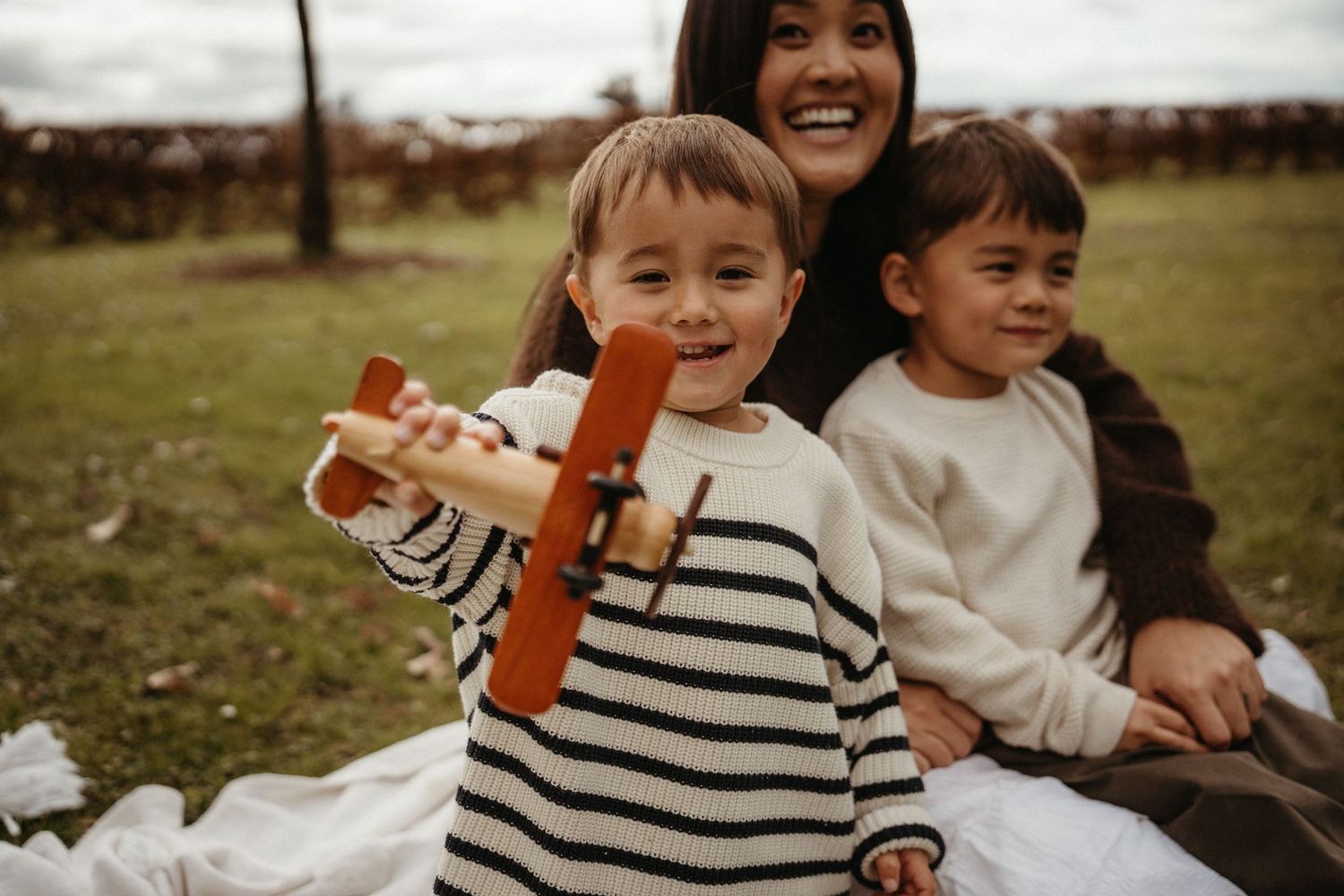 A young boy holds a wooden toy airplane while sitting with his mother and sibling on a blanket outdoors.