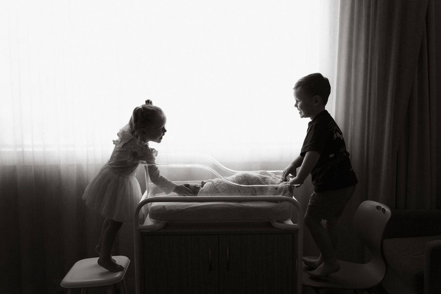 Black and white photo of two young children standing over a baby crib by a bright window.
