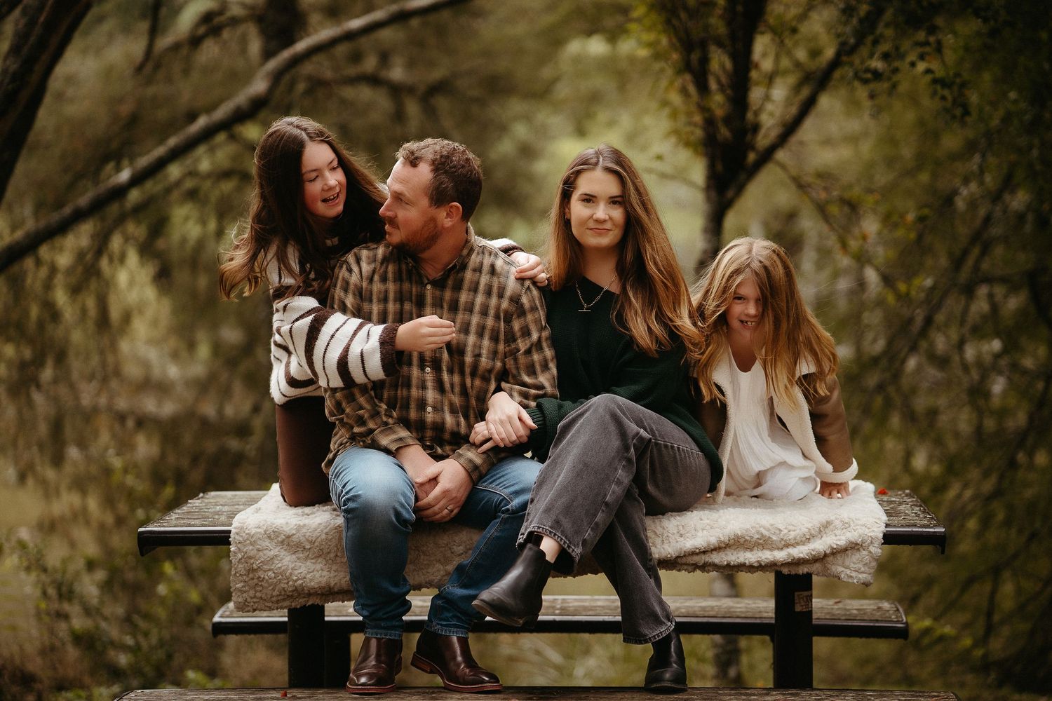 Family of four sitting on a picnic table bench laughing together in a wooded park setting.