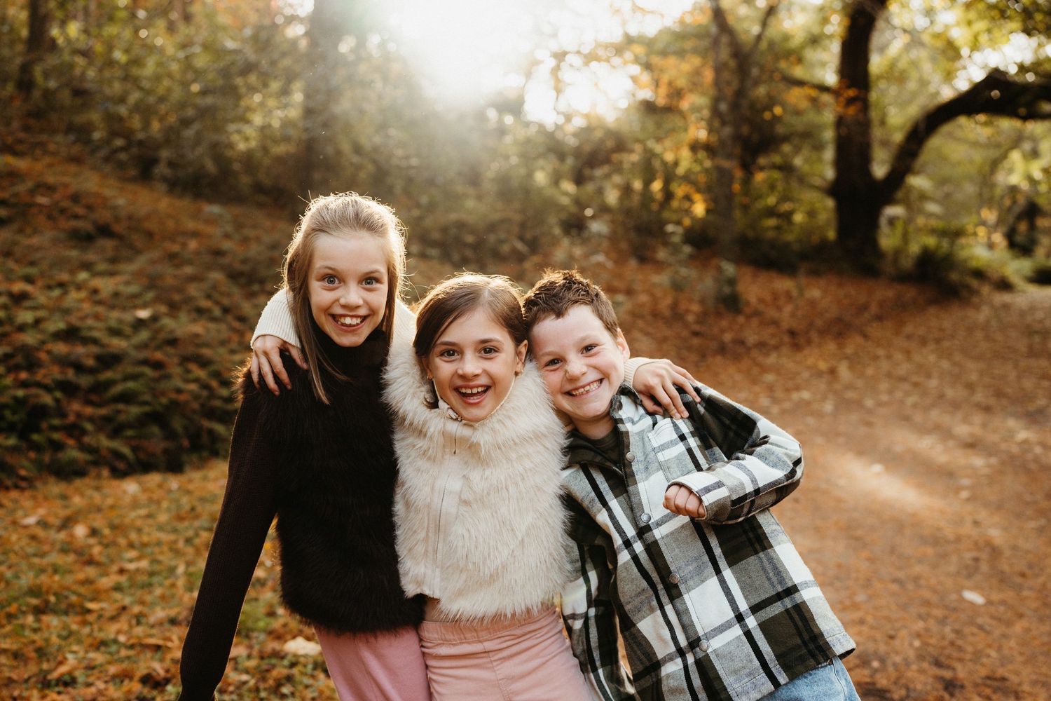 Three happy children hugging outdoors on a sunlit autumn forest path.