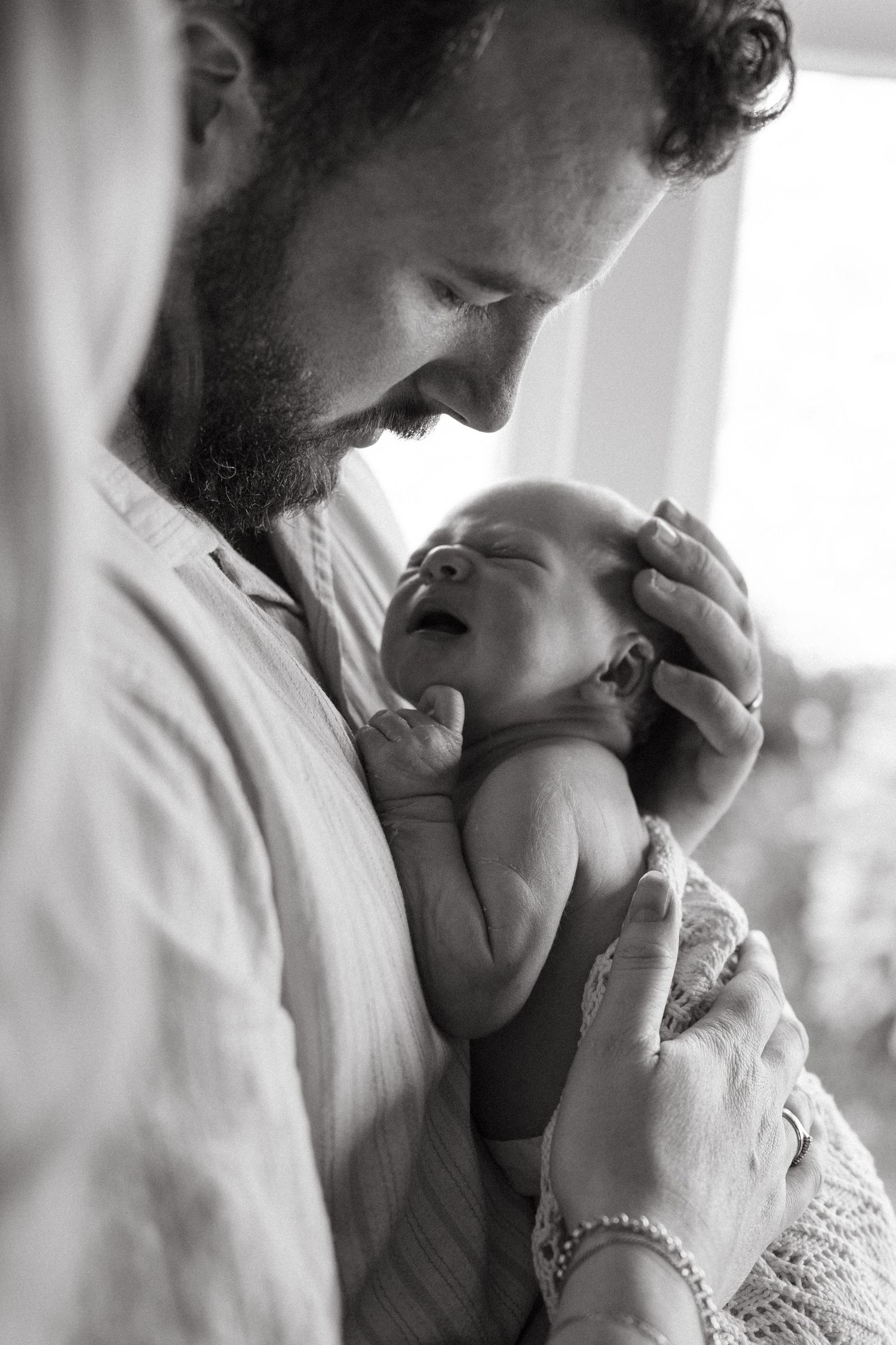 Black and white photo of a bearded father tenderly holding his newborn baby by a window.