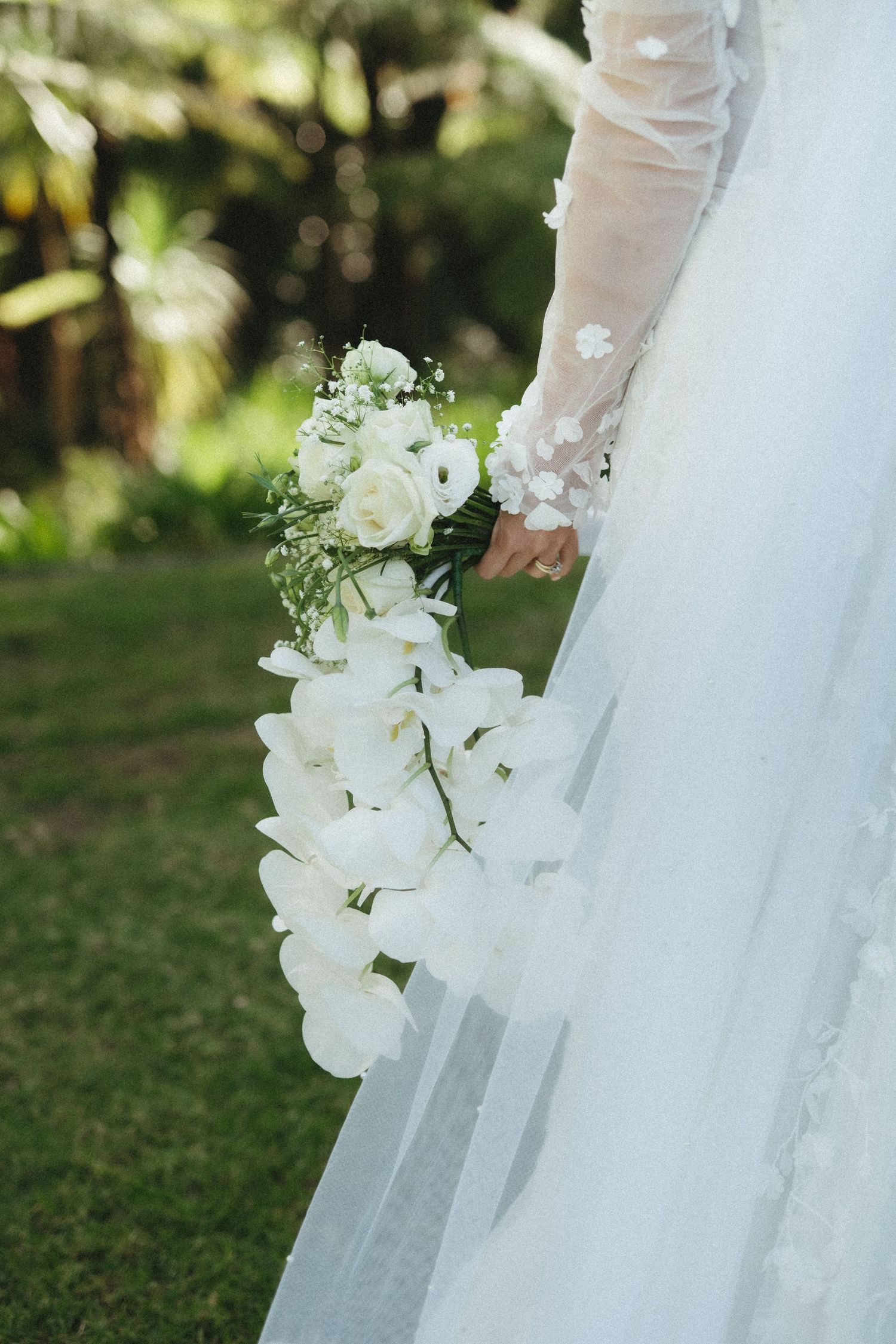 Bride holding a cascading white orchid and rose wedding bouquet against a lush green background.