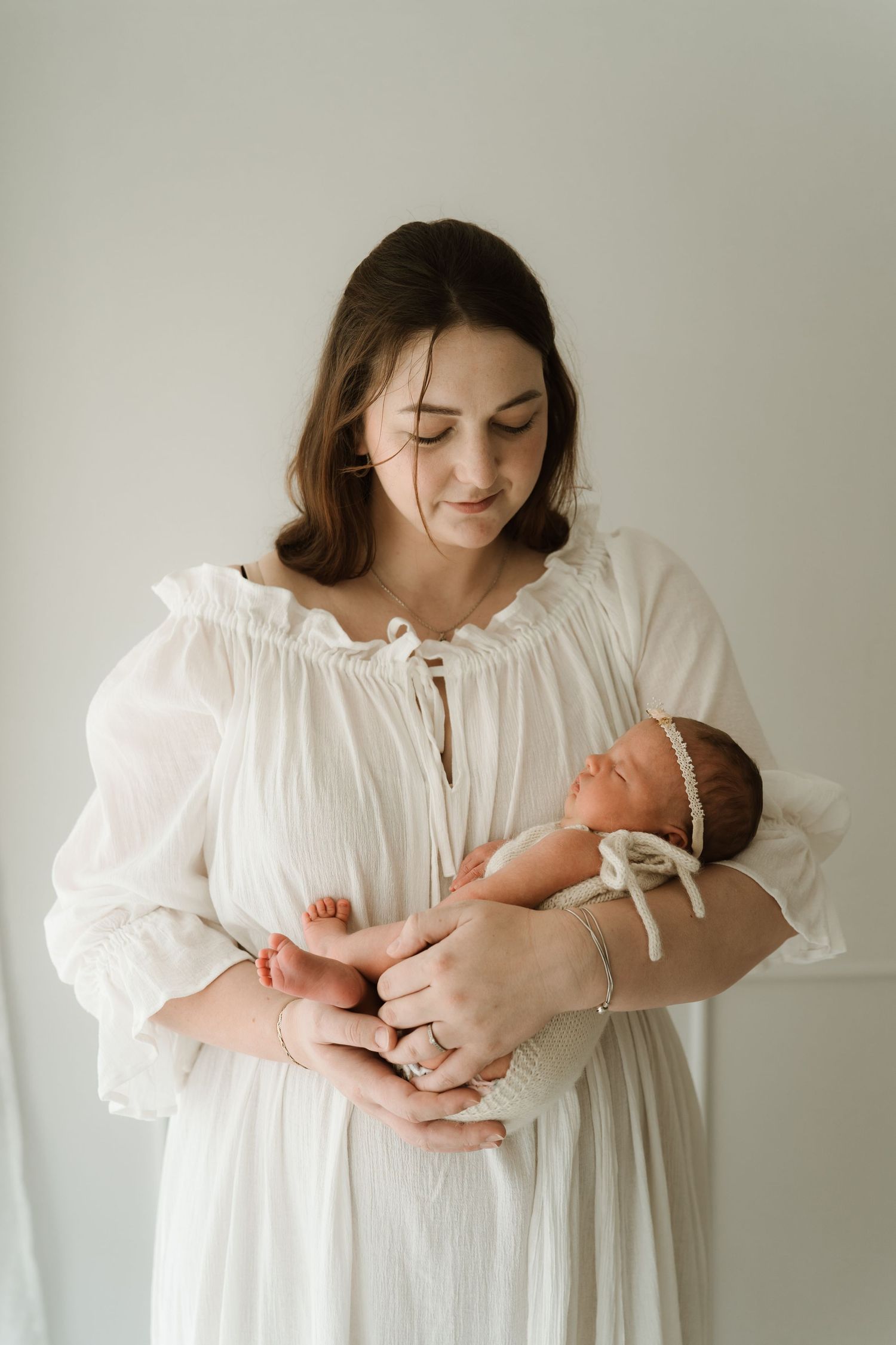 A mother in a white dress tenderly holds her sleeping newborn baby, wearing a delicate headband, against a soft background.