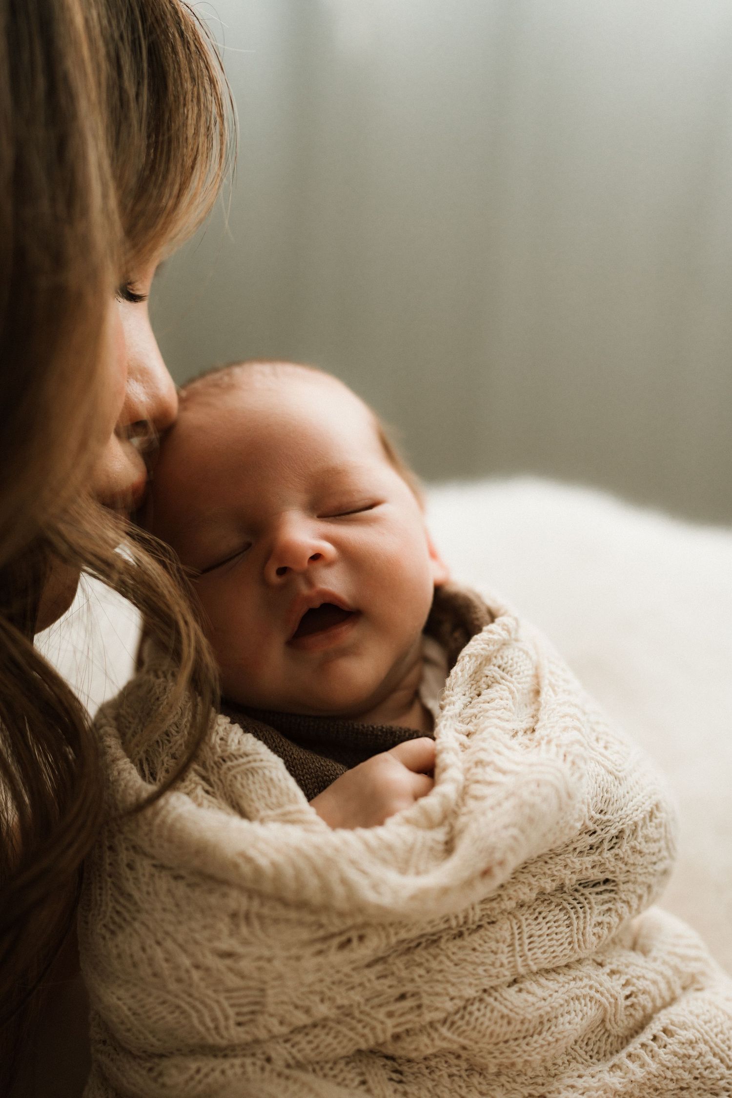 A mother tenderly kisses her sleeping newborn baby wrapped in a cozy cream knit blanket.