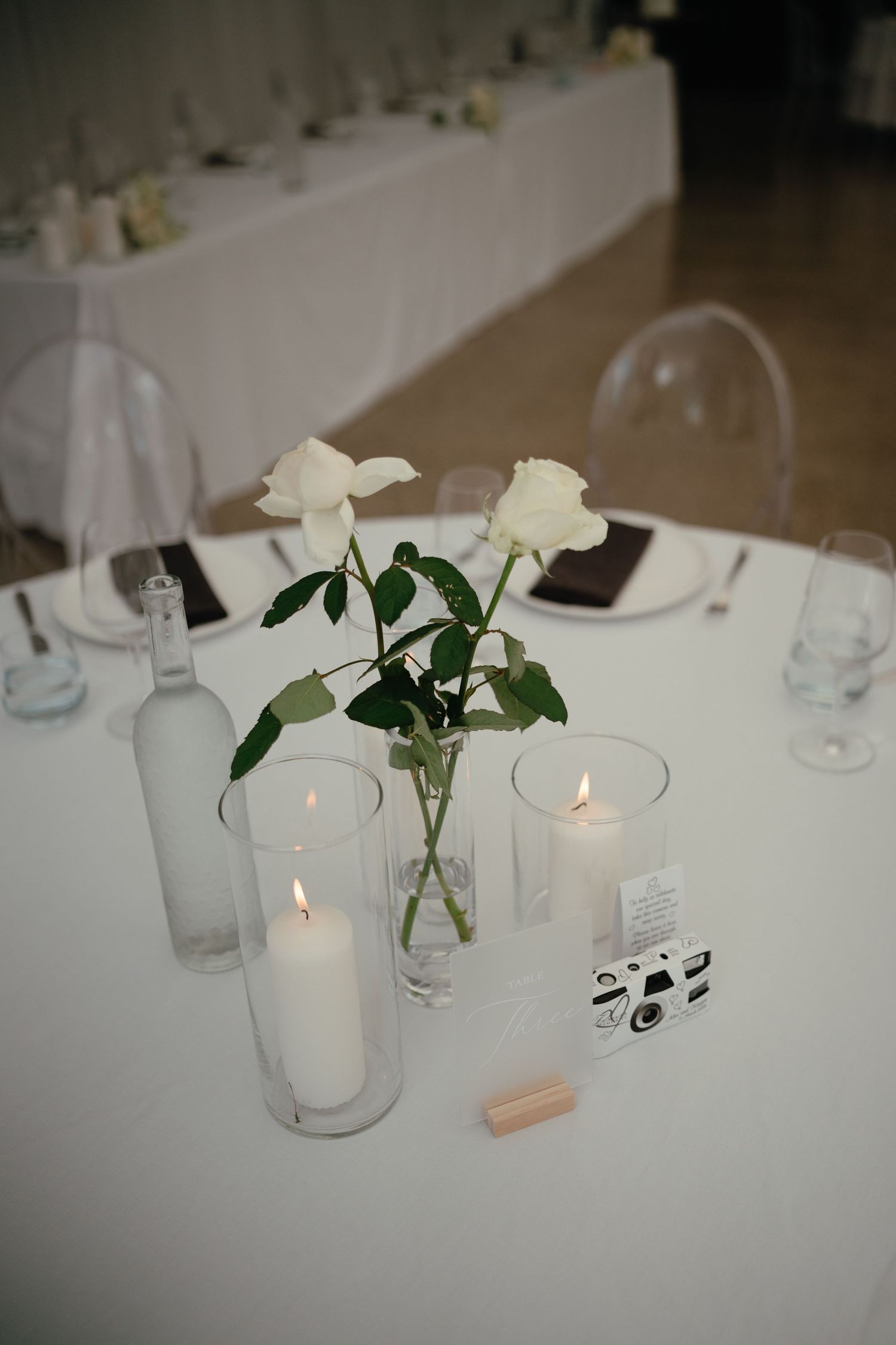 Minimalist wedding reception table with white roses, pillar candles, and disposable camera favor.