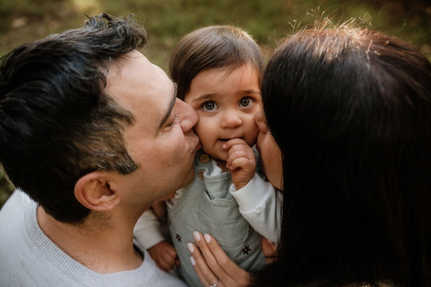 Loving parents kissing their wide-eyed baby on both cheeks simultaneously in outdoor setting.