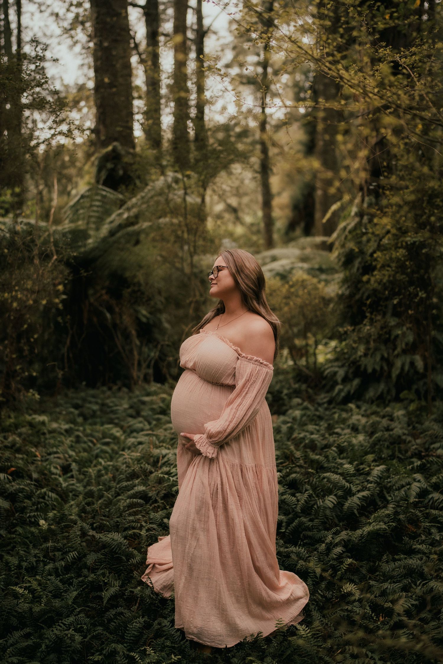 Pregnant woman in pink off-shoulder gown standing among lush ferns in a forest for a maternity photoshoot.