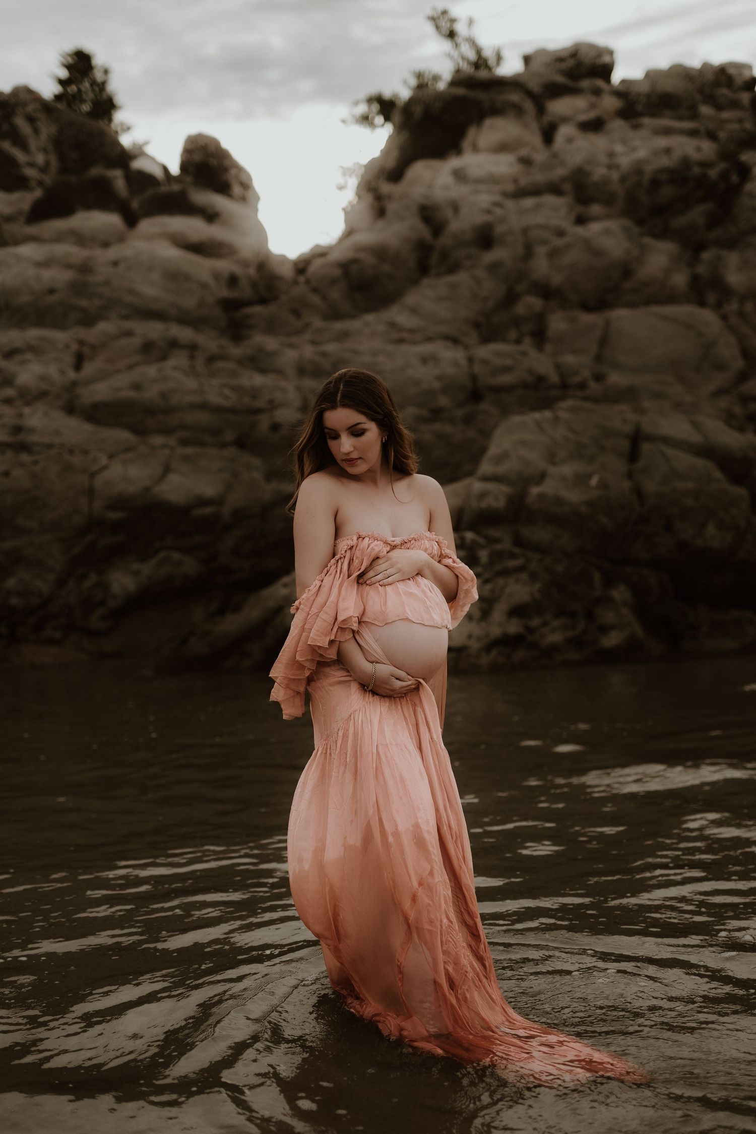 Pregnant woman in pink flowing gown standing in water with rocky backdrop during maternity photoshoot.