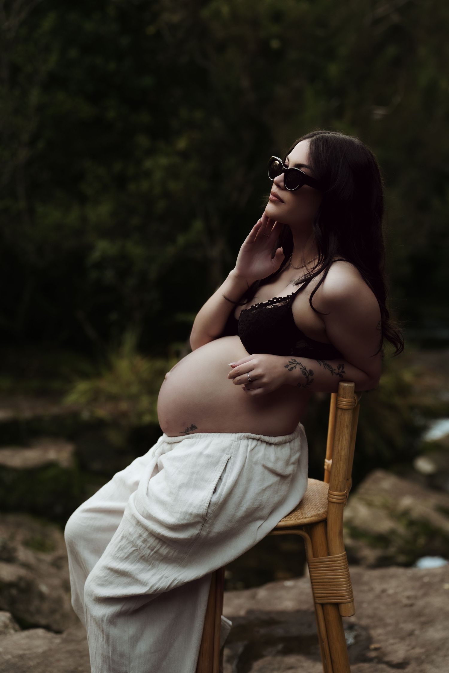 Pregnant woman in black lace bralette and white pants sits on chair outdoors, wearing sunglasses, cradling baby bump.