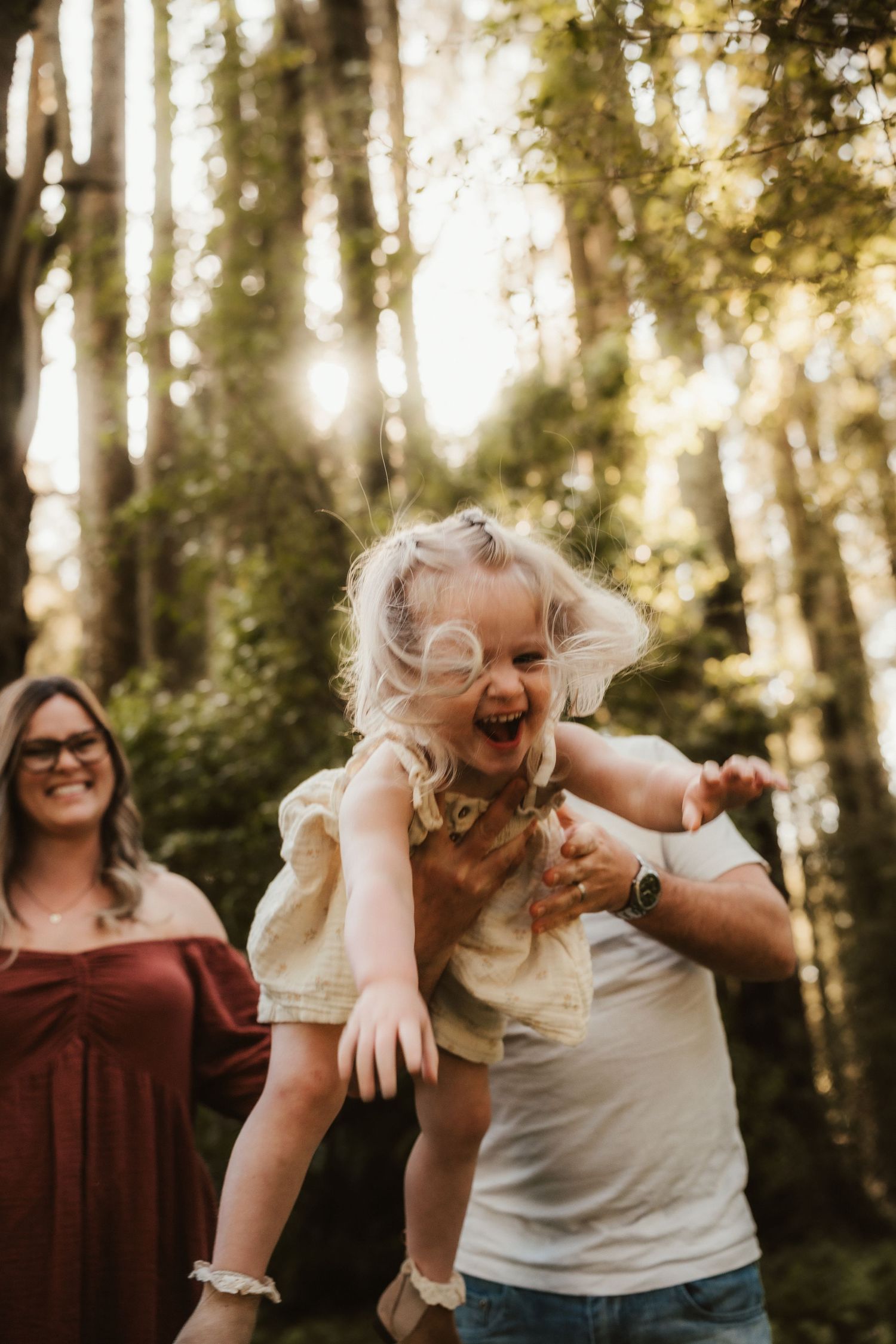 A joyful toddler girl laughs while being lifted by her father in a sunlit forest, with her smiling mother nearby.