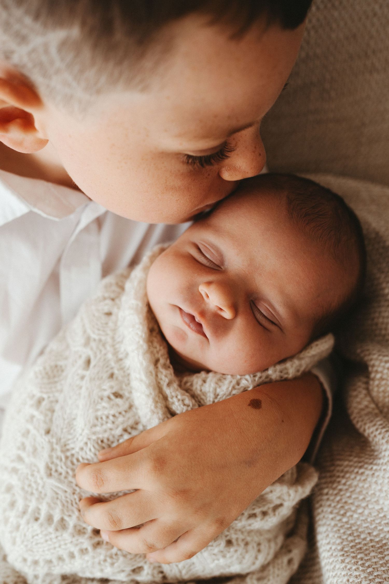 Older child gently kissing sleeping newborn baby wrapped in a soft cream knit blanket.