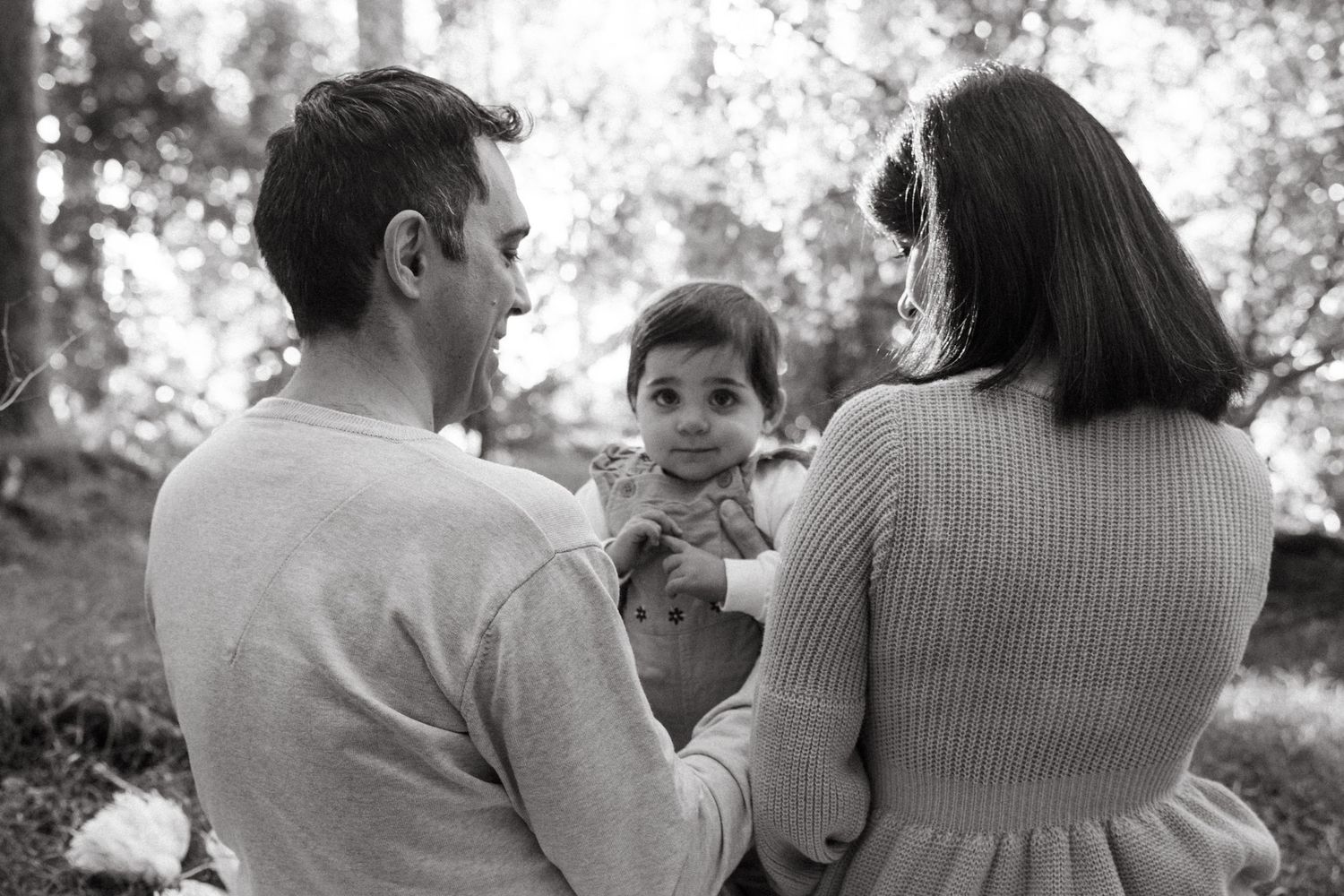 Black and white photo of parents holding smiling toddler facing away in a wooded park setting.
