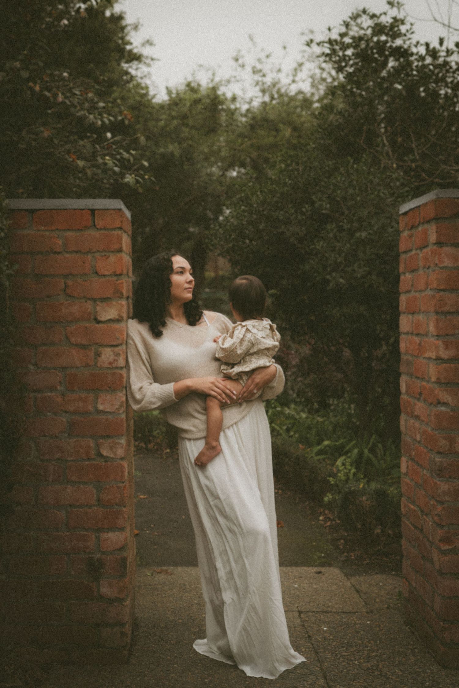 A woman in a white gown holds a baby between brick pillars surrounded by lush greenery.