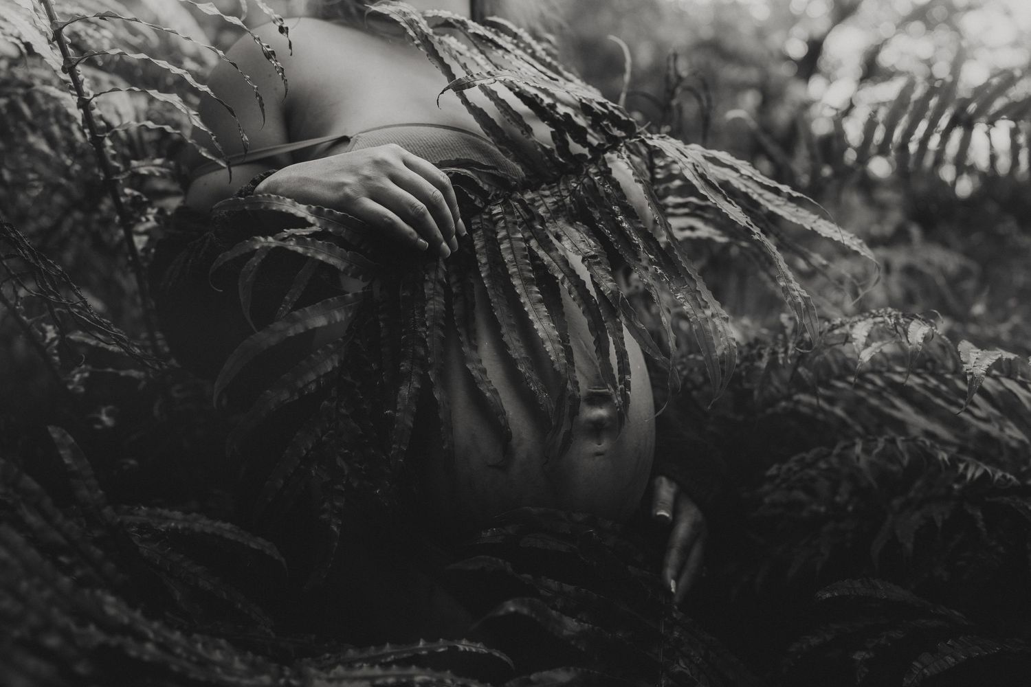 Black and white photo of a person lying among lush fern leaves in a forest setting.