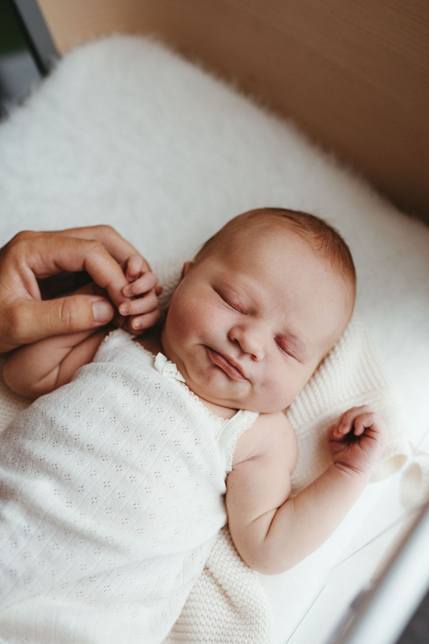 Sleeping newborn baby smiling while an adult gently holds their tiny hand on a white knit blanket.