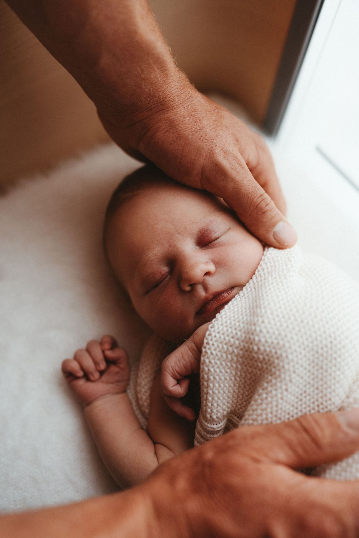 Sleeping newborn baby in white knit wrap with parents hands gently cradling head.