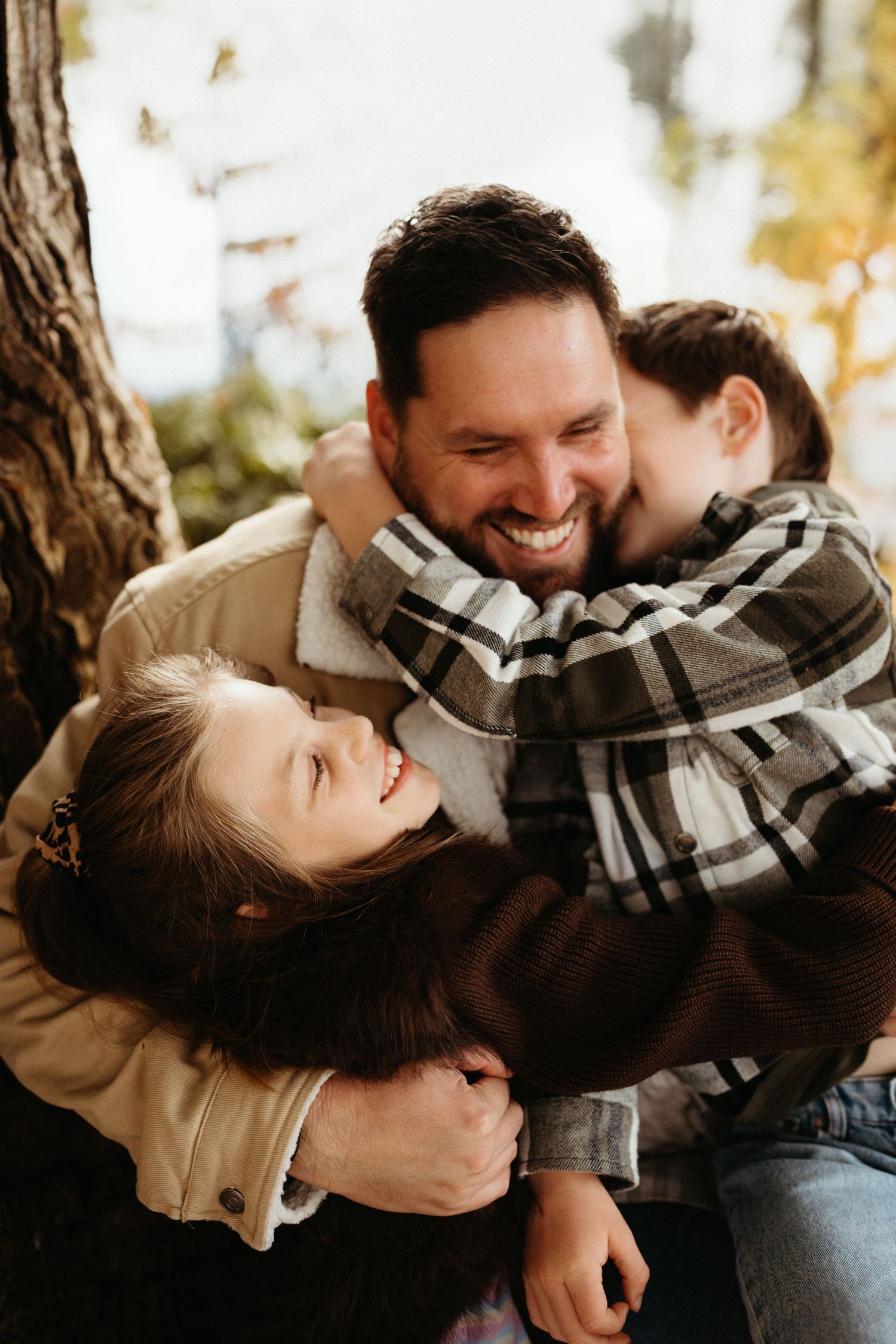 A smiling father hugs two laughing children outdoors near a tree in a warm autumn family portrait.