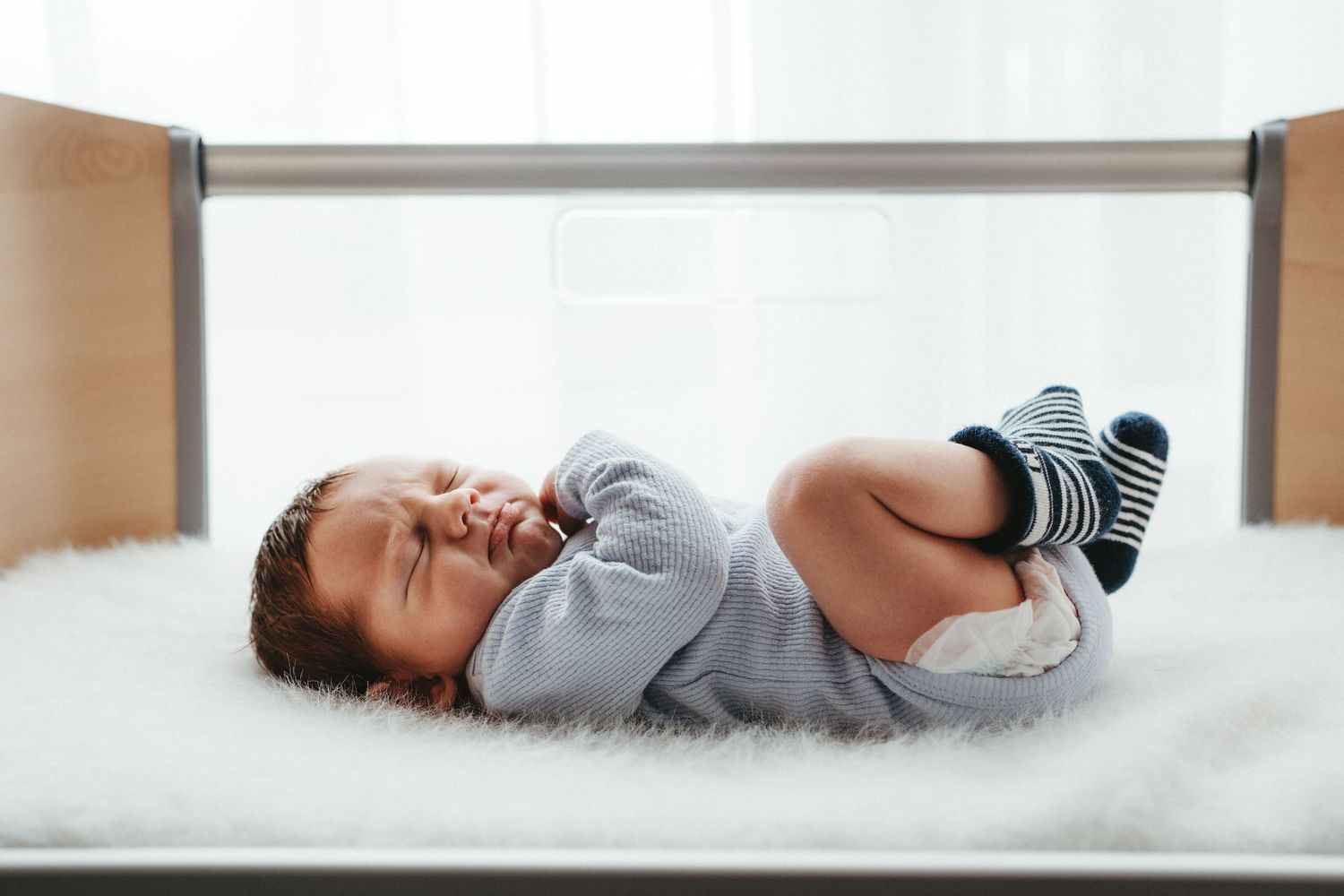 Cute baby in striped socks lying on a fluffy white blanket in a wooden crib.