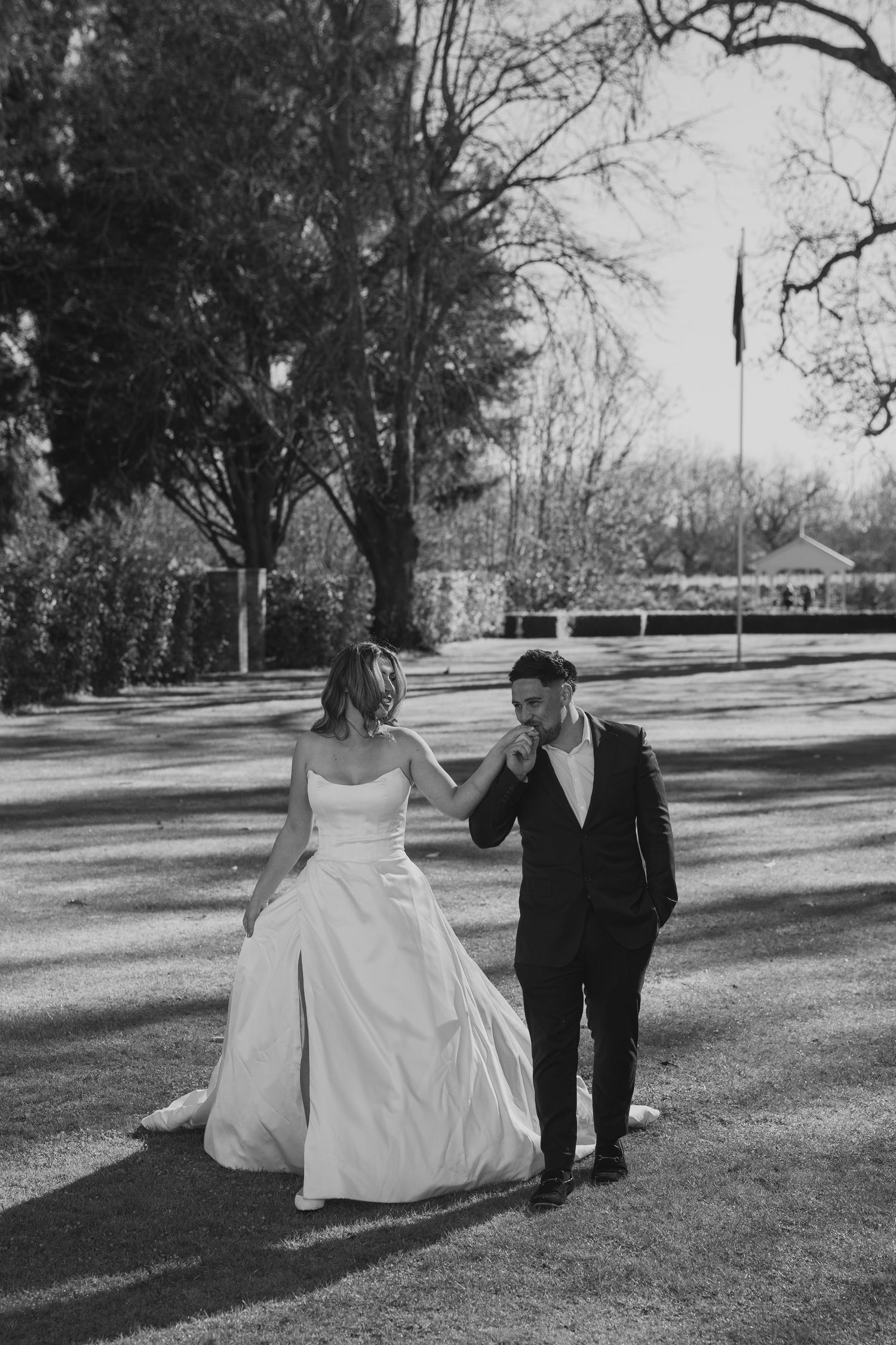 Groom kisses bride's hand as they walk together across sunlit garden grounds in black and white.