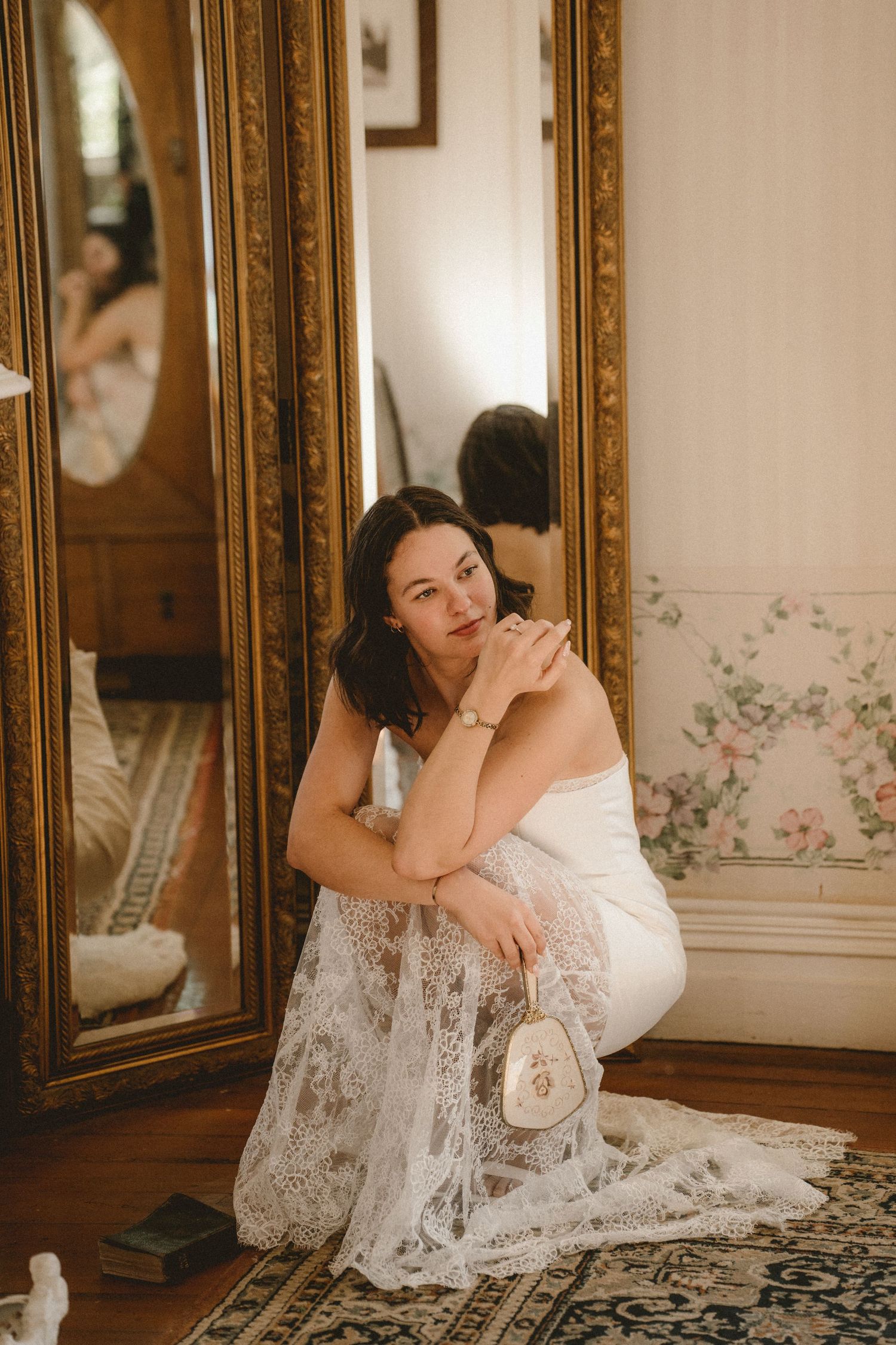 Bride in white lace wedding dress sits beside ornate gold mirrors in a vintage floral wallpapered room.