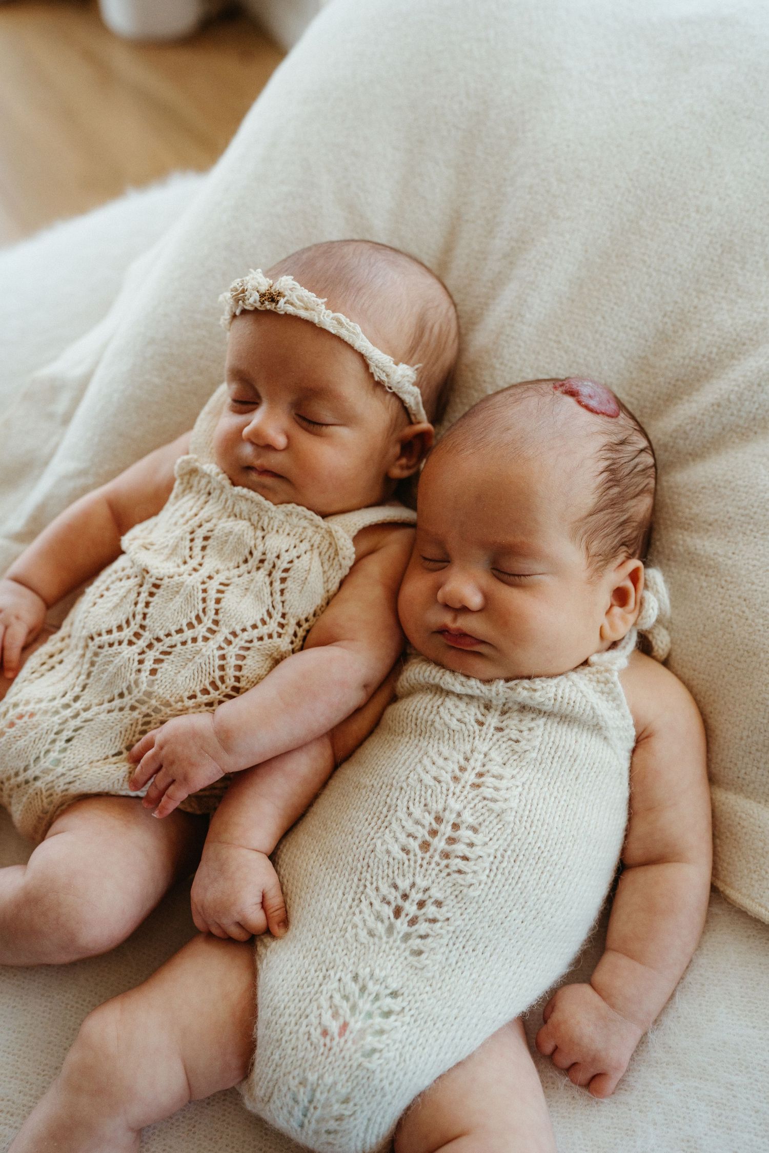 Two sleeping newborn twins in cream knit rompers rest together on a soft white cushion.