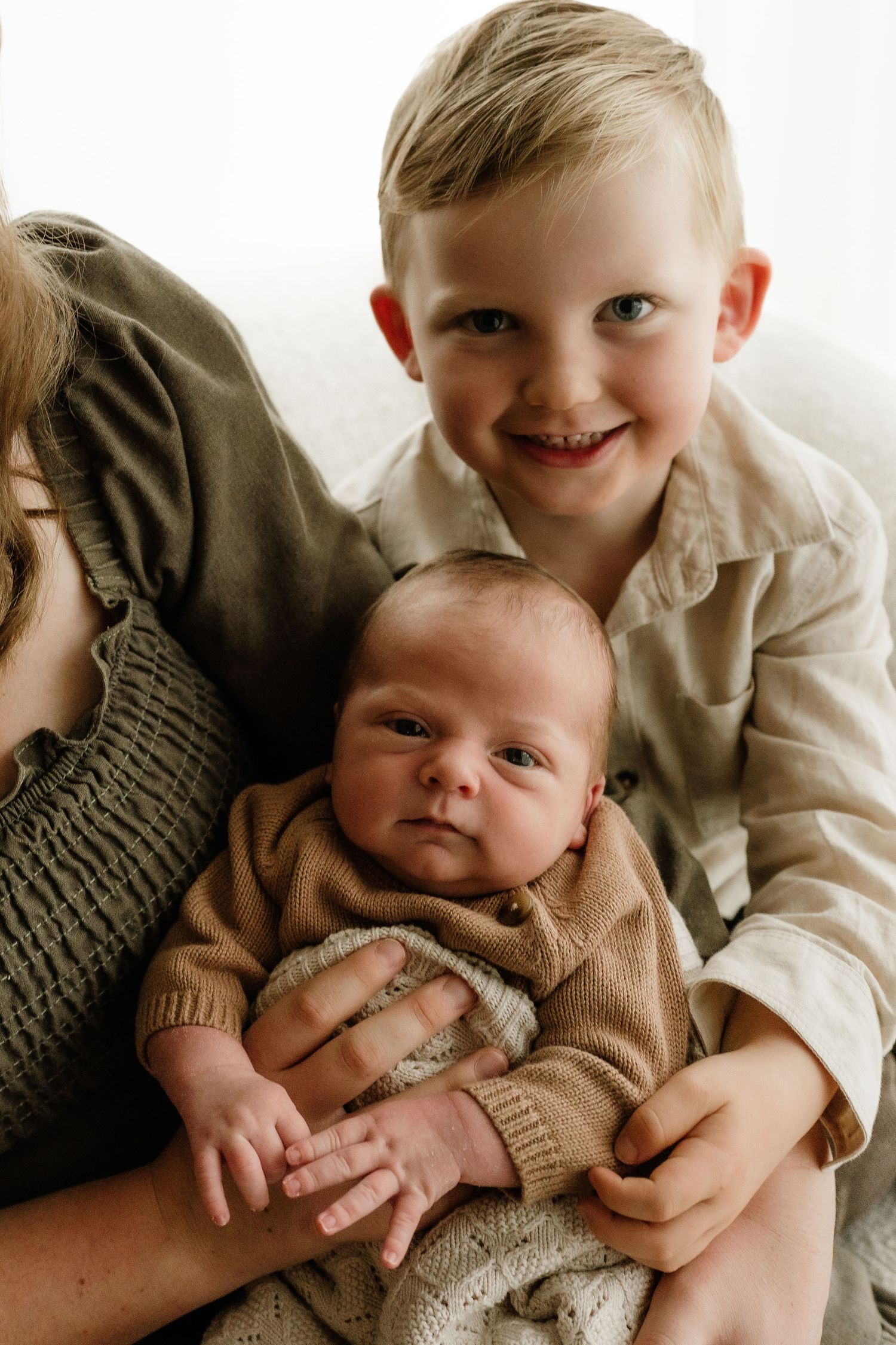 A smiling young boy poses with a newborn baby in a knit outfit, held by a woman in a soft, natural-light setting.