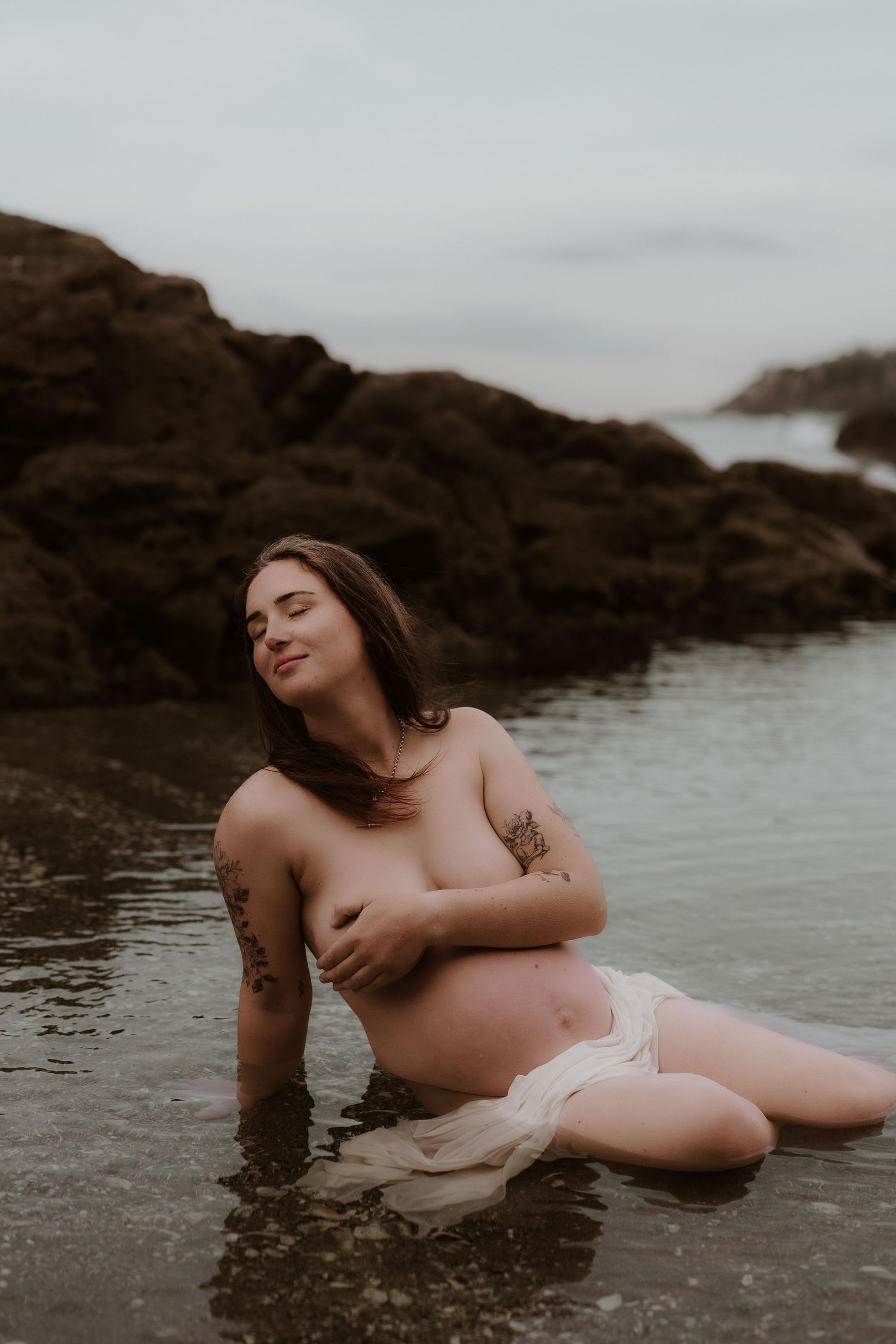 Pregnant woman sitting in shallow ocean water near rocks, cradling her baby bump during a maternity photo shoot.