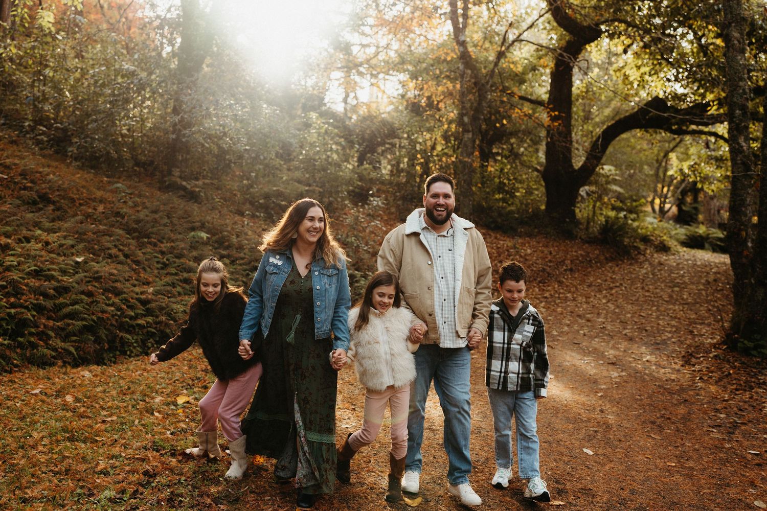 Family of five walking on autumn forest path, smiling, surrounded by golden fall foliage and warm sunlight.