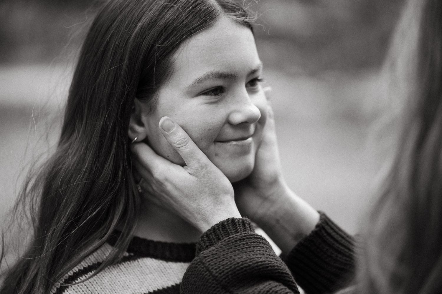 Black and white photo of a smiling girl having her face gently held by someone's hands outdoors.