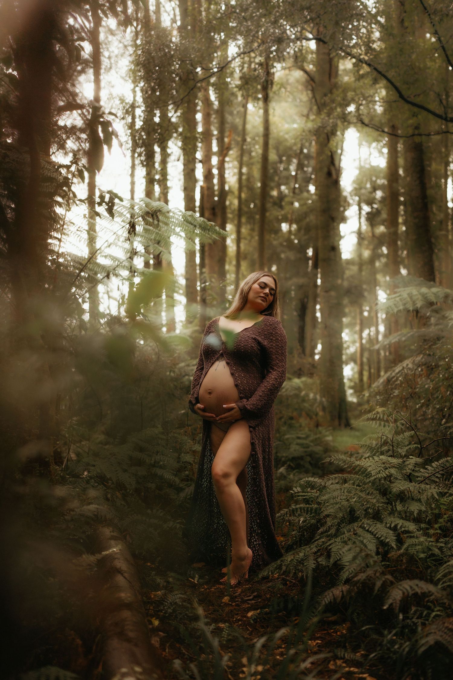Pregnant woman in sheer mauve gown cradling baby bump among lush forest ferns in soft natural light.