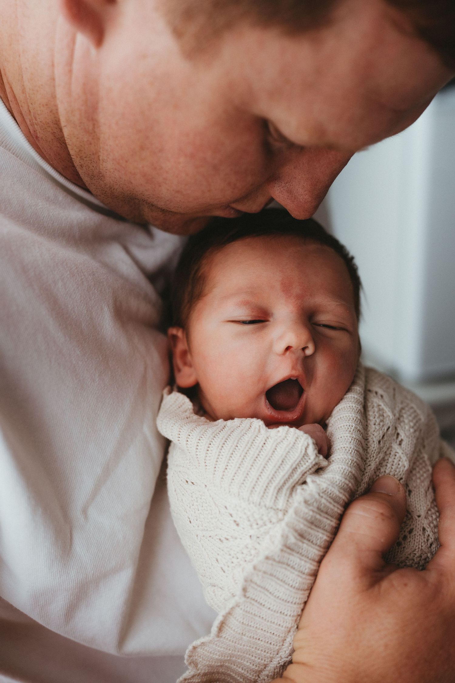 Father tenderly holding a yawning newborn baby wrapped in a knit blanket.