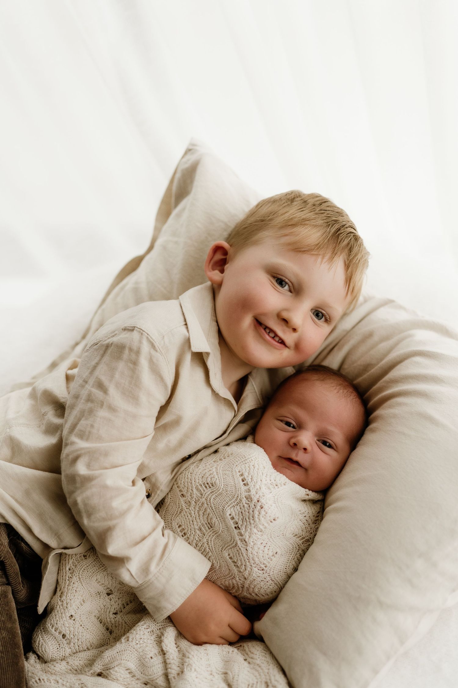 Smiling blonde toddler boy gently holding a swaddled newborn baby on a white pillow backdrop.